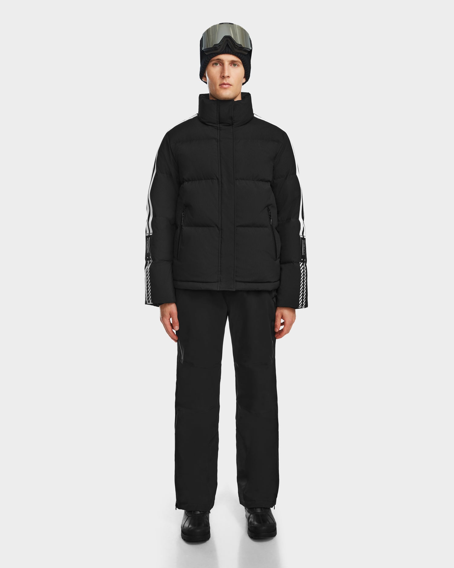 A person wearing the Rudsak USHER - 6124516 Black water-resistant ski jacket and pants, black boots, a black beanie, and ski goggles stands facing forward against a plain white background.