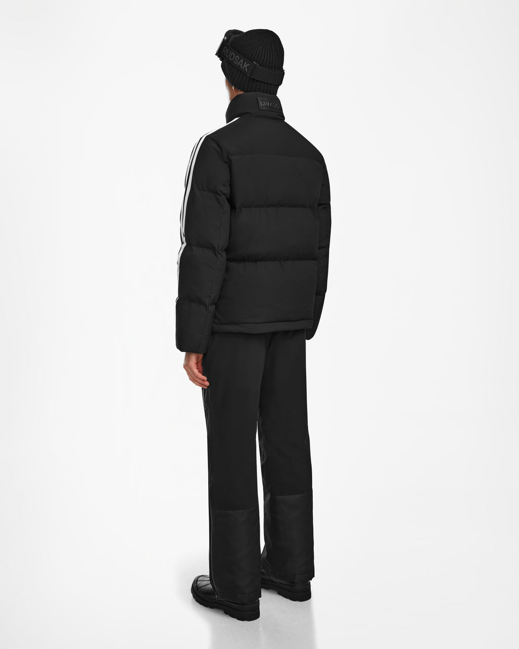 A person faces away, wearing the Rudsak USHER - 6124516 Black RDS-certified down jacket with white sleeve stripes, black pants, shoes, and beanie hat. The background is plain white.