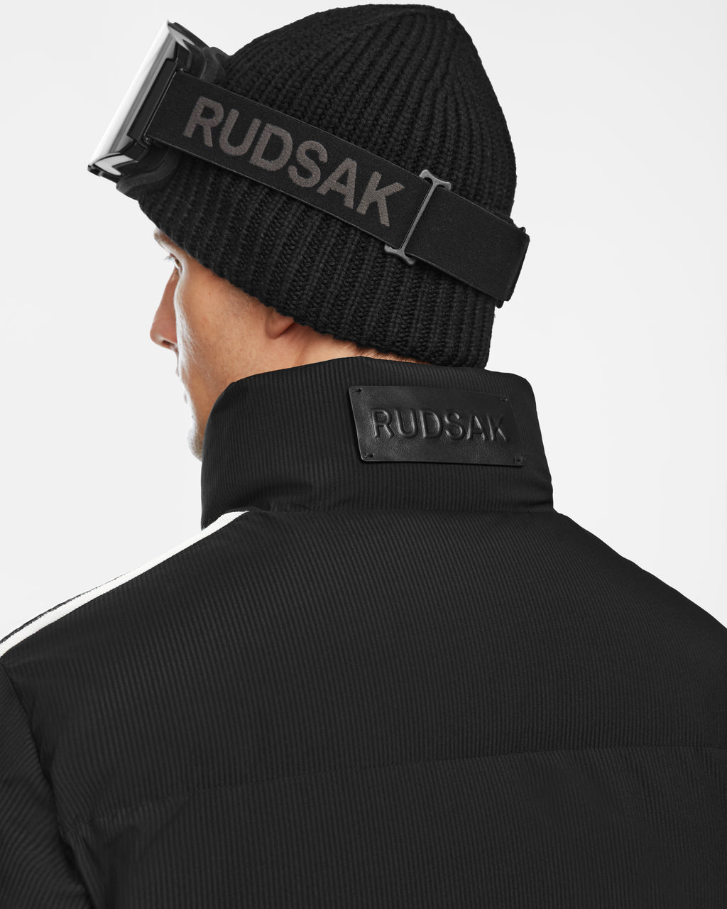 A person is shown from the back wearing the Rudsak USHER - 6124516 Black men’s water-resistant down ski jacket, paired with a black knit beanie and Rudsak-branded black ski goggles.