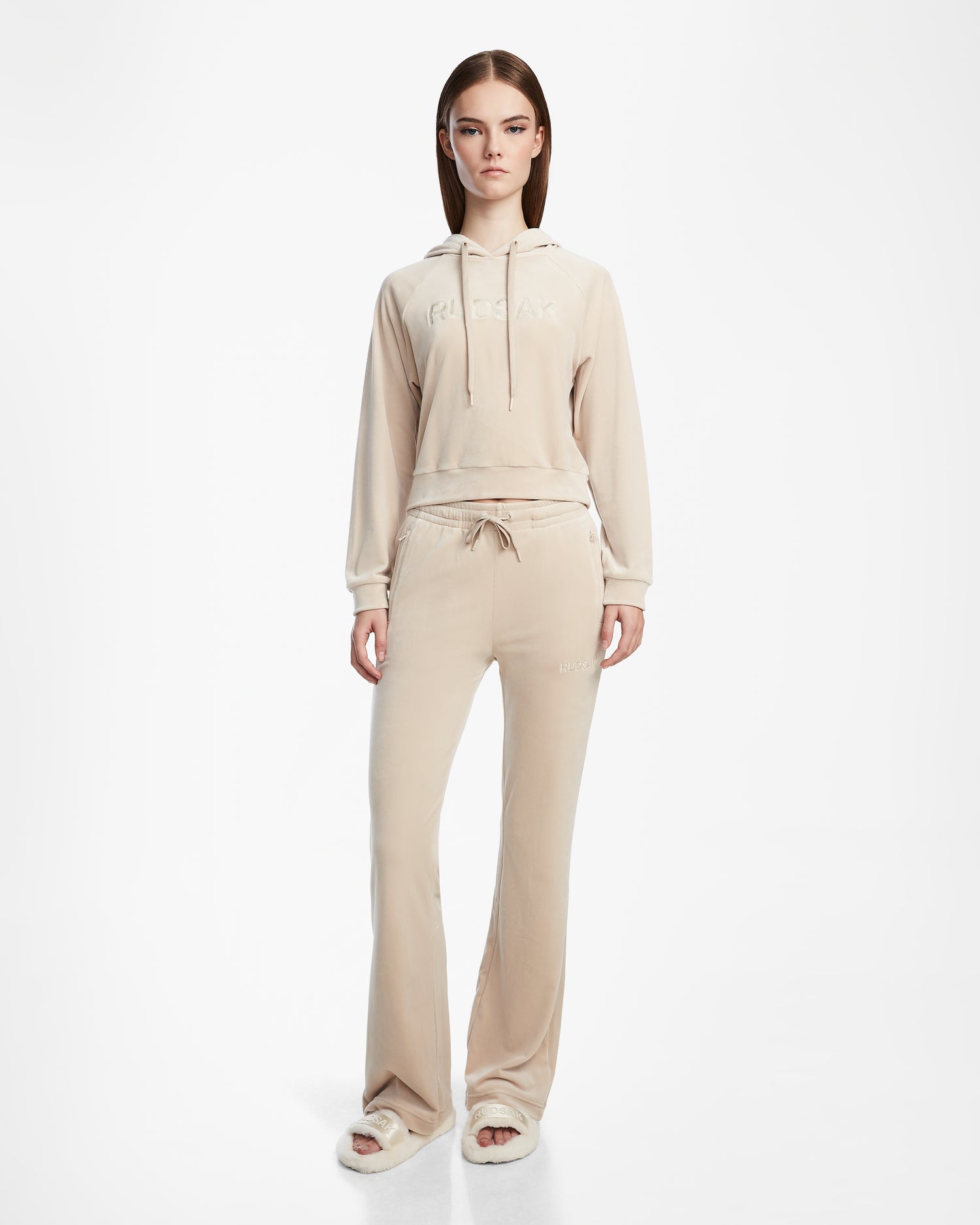 A woman stands facing forward, wearing the Rudsak VALERIA - L523500 Cream velour hoodie and matching drawstring pants with open-toe slippers, set against a plain white background.