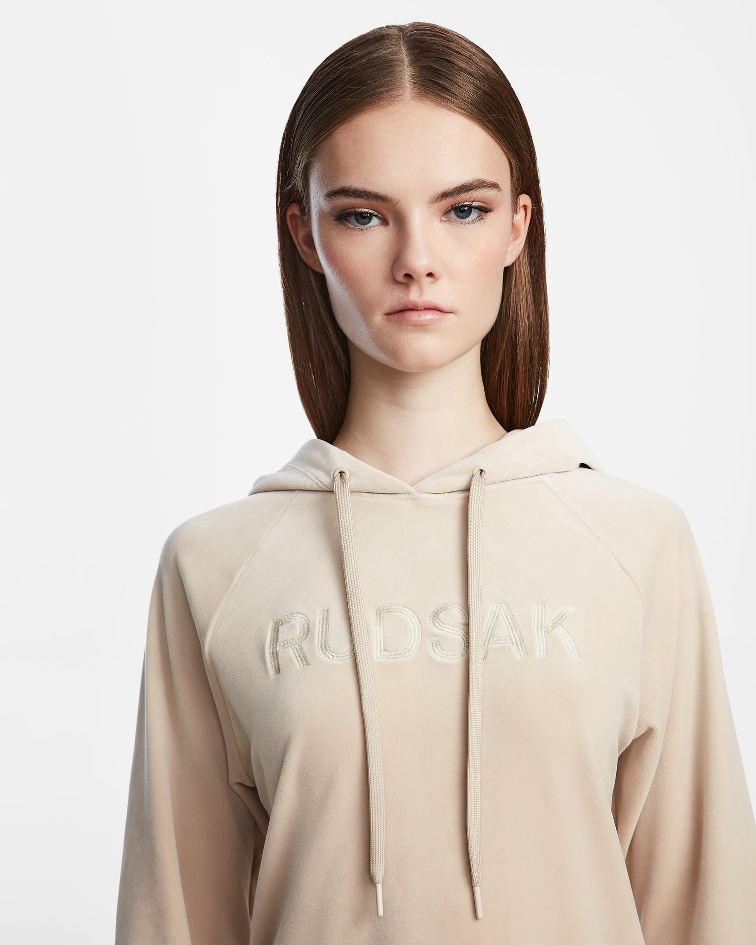 A young woman with straight brown hair wears the Rudsak VALERIA - L523500 Cream hoodie with RUDSAK embroidery on the front. She has a neutral expression and stands against a plain white background.