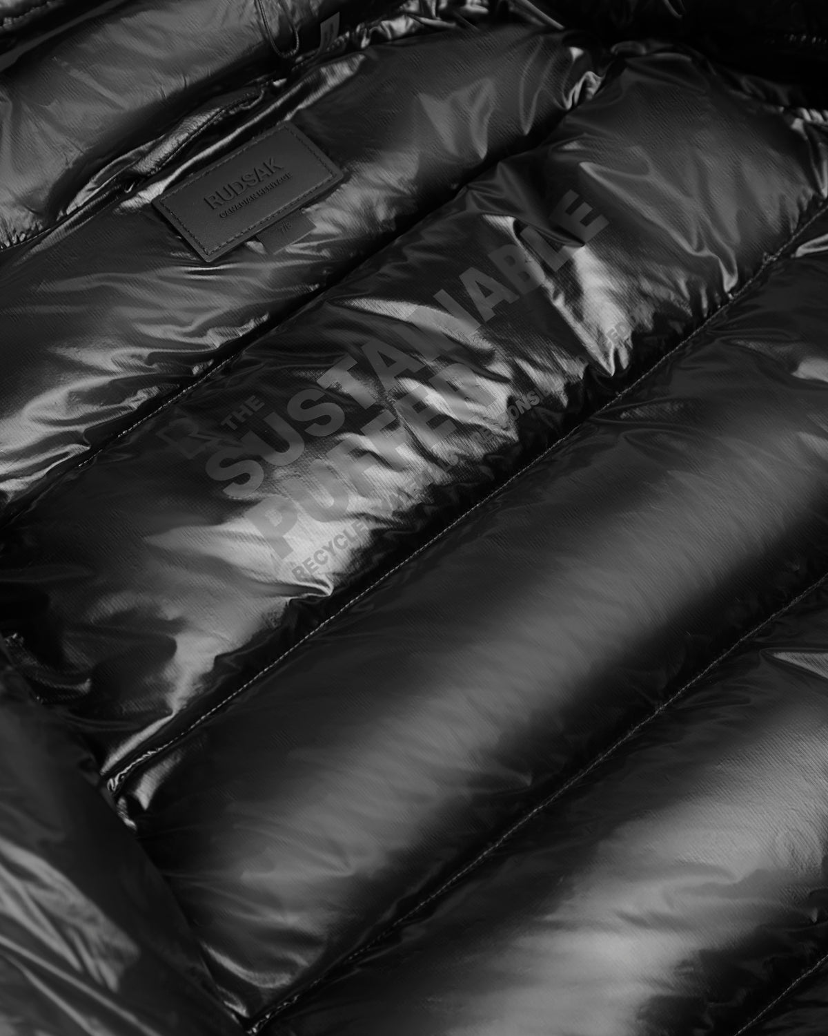 Close-up of the VINCE KS - 0124502 Black unisex winter jacket by Rudsak, featuring partial “THE SUSTAINABLE FUTURE” and “RECYCLED” text, a Rudsak label near the top, and crafted with RDS-certified down for warmth and style.