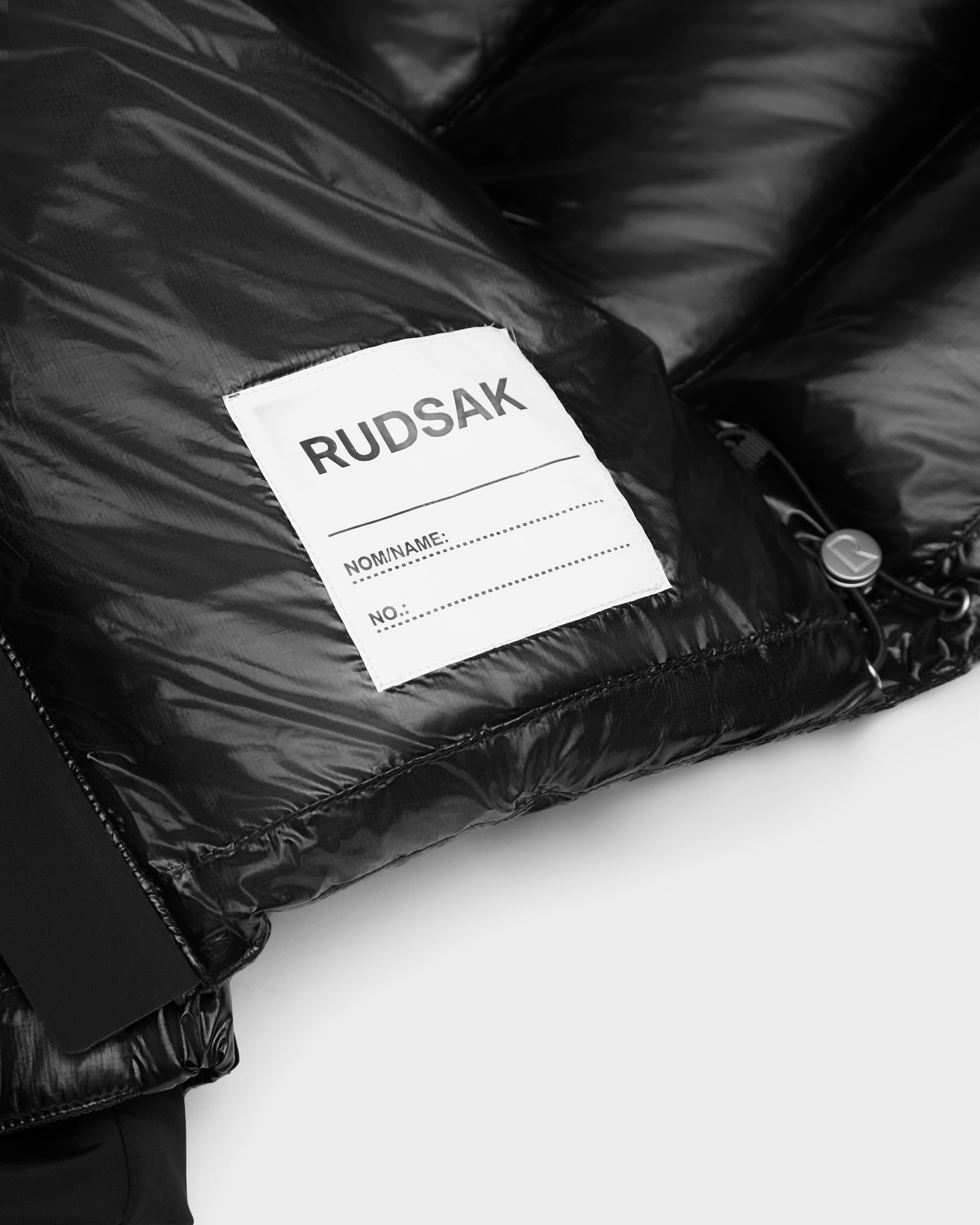 A close-up of the Rudsak VINCE KS - 0124502 Black kids’ unisex down puffer jacket shows its white label with blank lines for Name and No., shiny fabric, zipper detail, and RDS-certified down filling for warmth.