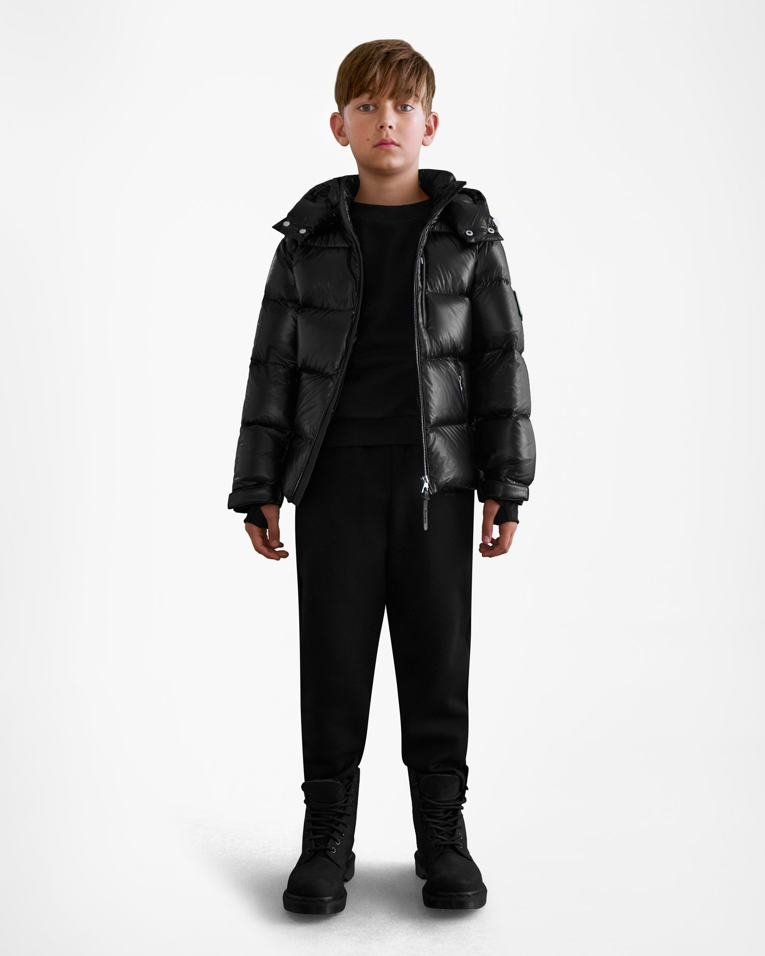 A young boy, facing forward against a white background, wears the Rudsak VINCE KS - 0124502 Black unisex winter jacket with matching black pants and boots.