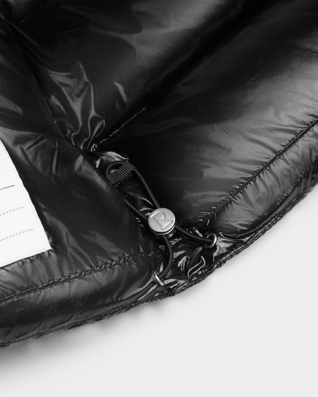 Close-up of the VINCE KS - 0124502 Black unisex winter jacket by Rudsak, featuring a shiny finish, drawstring and toggle-adjustable hem, RDS-certified down filling, and a small white tag on the left side.