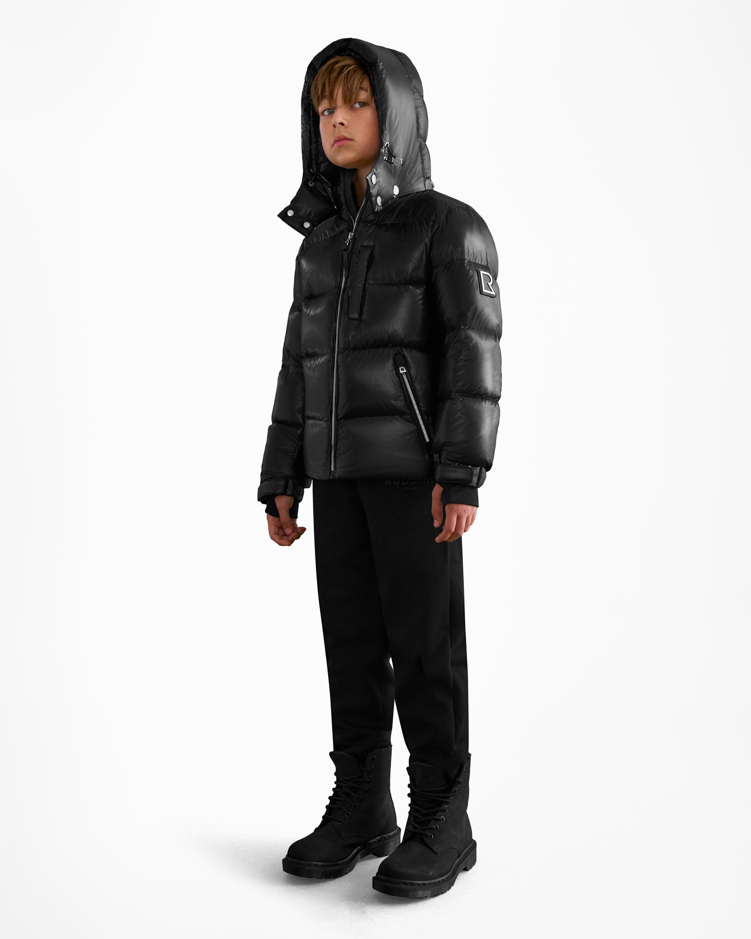 A child stands against a plain white background, wearing the Rudsak VINCE KS - 0124502 Black unisex RDS-certified down puffer jacket with zippered pockets, a hood, and sleeve patch—paired with black pants and boots for a cozy, stylish look.