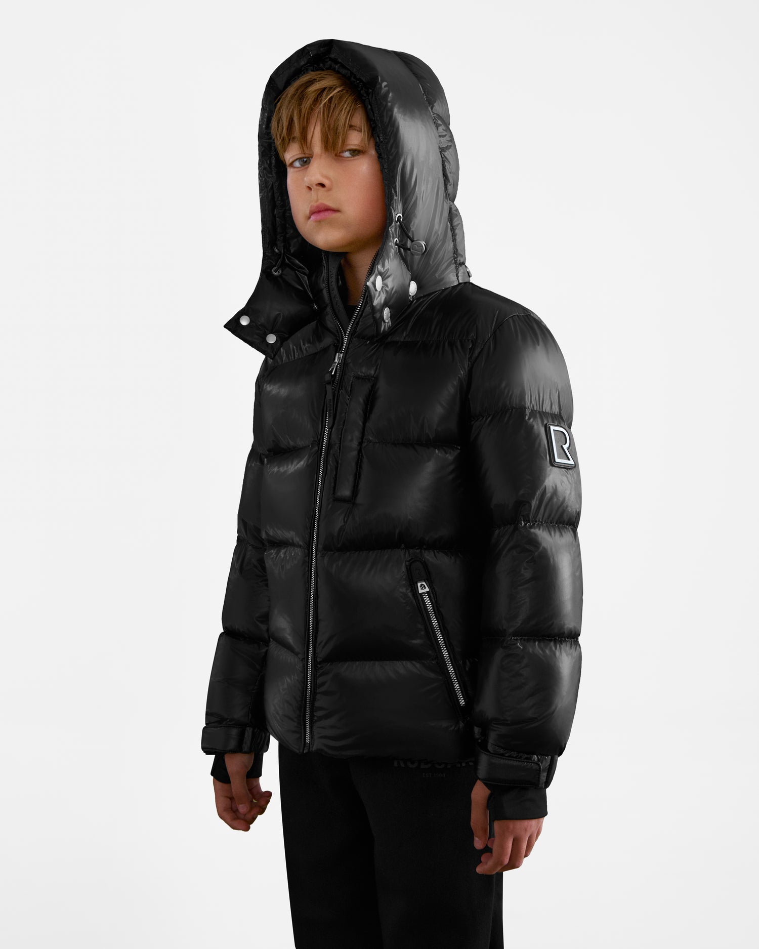 A young boy with blond hair wears the Rudsak VINCE KS - 0124502 Black kids down puffer jacket with the hood up, standing slightly to the side and looking at the camera against a plain white background.