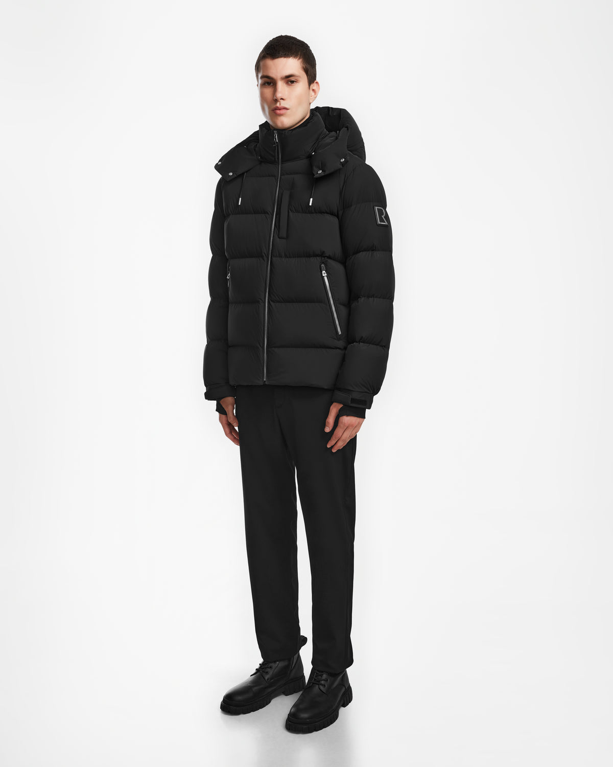 A person stands against a plain white background wearing the Rudsak VINCE M - 6124561 Black men’s down puffer jacket with silver zippers, paired with black trousers and dress shoes—a sleek, weather-resistant monochrome look.