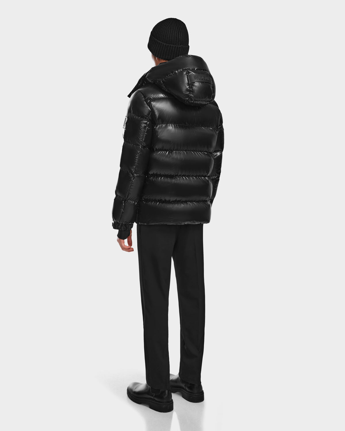 A person faces away, wearing the Rudsak VINCE S - 6124560 Black mens down puffer jacket with a hood, black pants, shoes, and a knit beanie, set against a plain white background.