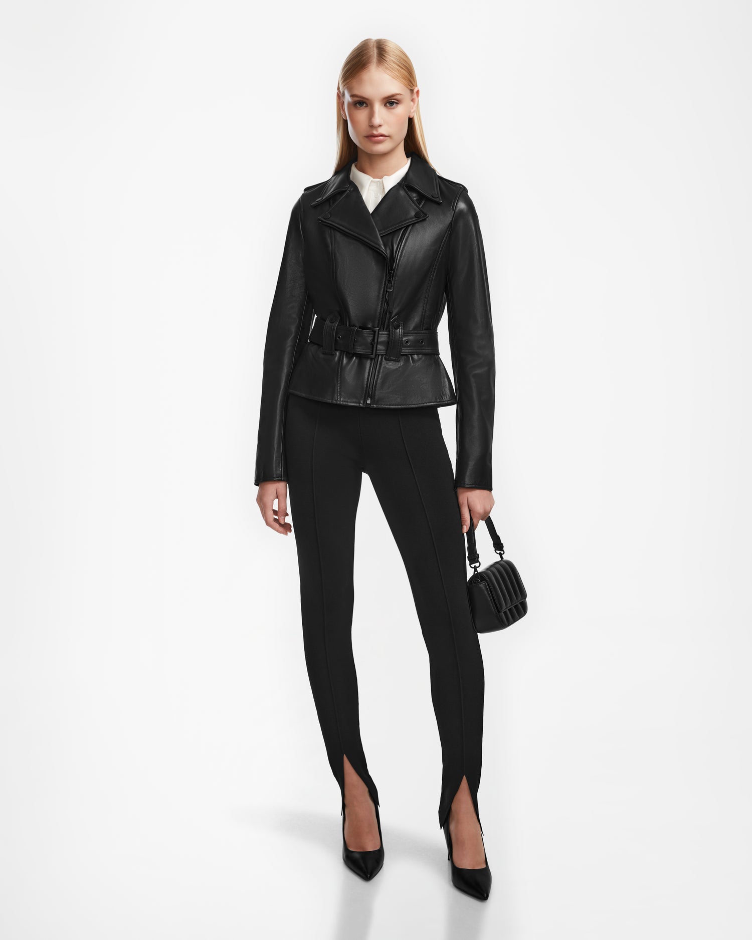 A woman stands against a white background wearing the Rudsak YEVA - 8124511 Black leather jacket, black slim-fit pants with ankle slits, black pointed heels, and holding a small black handbag.