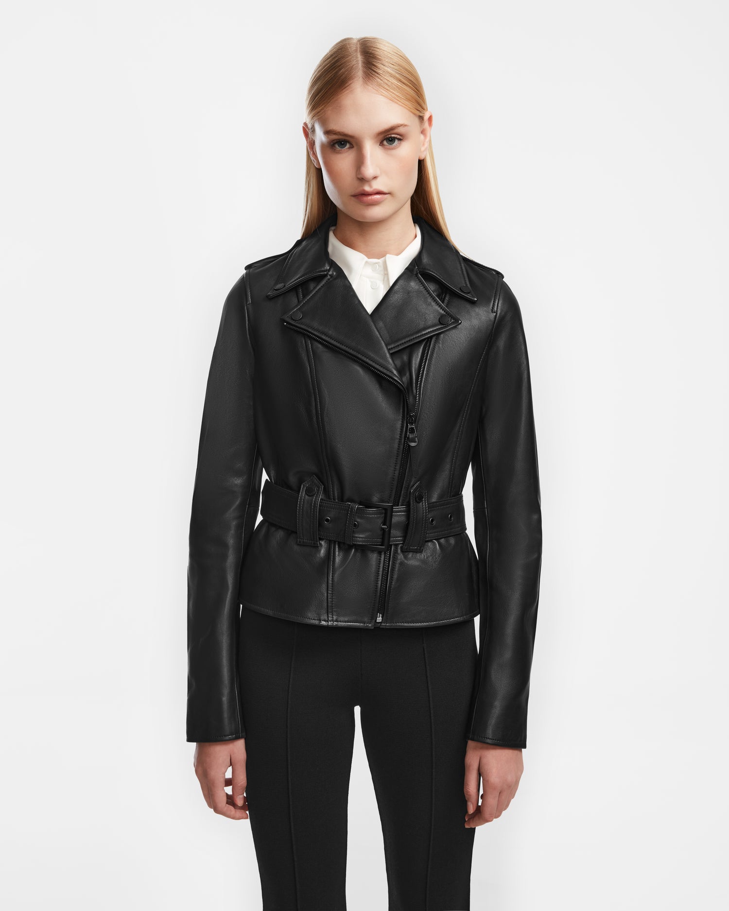 A woman with straight blonde hair wears the Rudsak YEVA - 8124511 black leather jacket over a white blouse and black pants, standing against a plain white background.