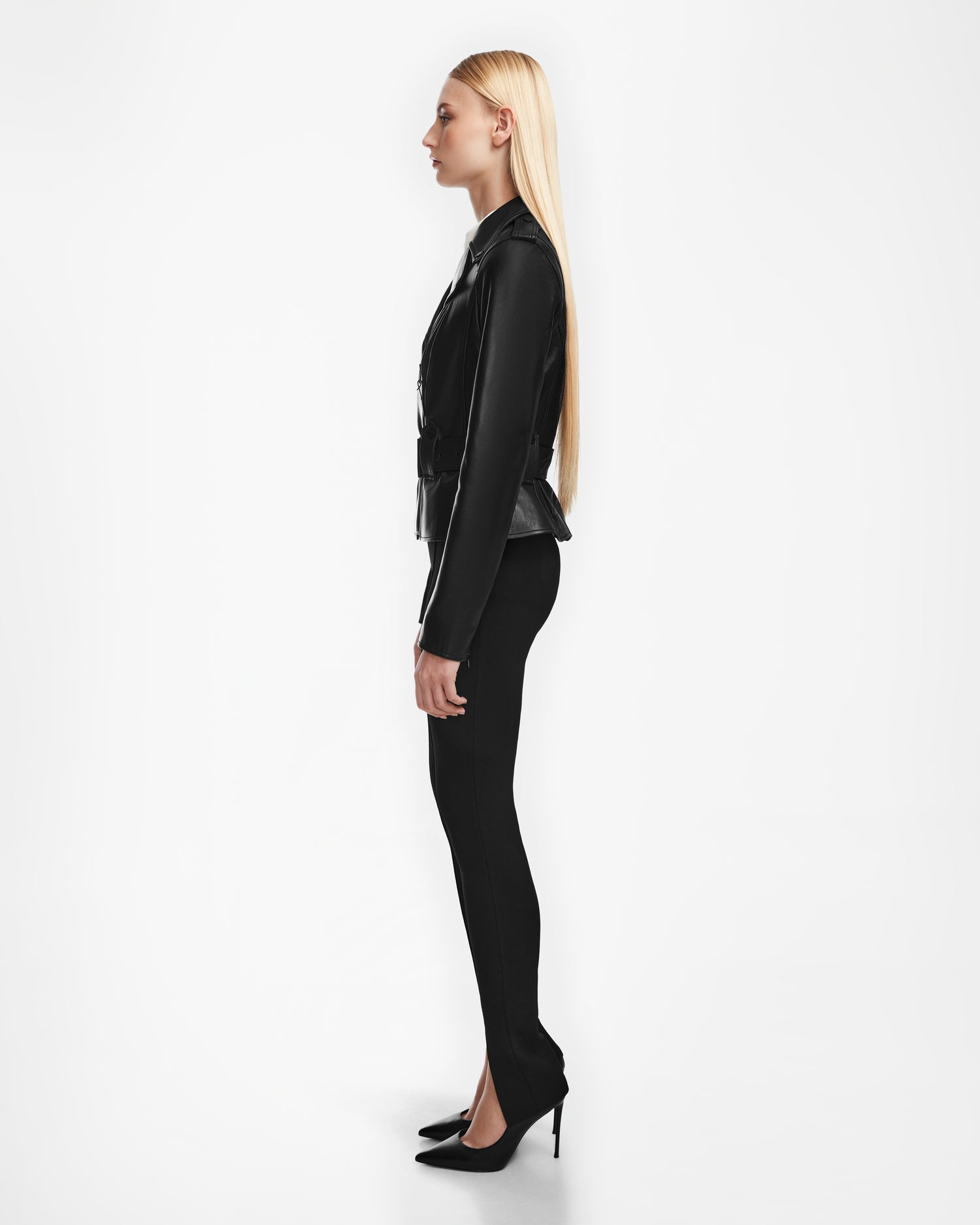 A woman with long blonde hair stands in profile against a white background, wearing the Rudsak YEVA - 8124511 Black leather jacket, black pants, and black high heels.