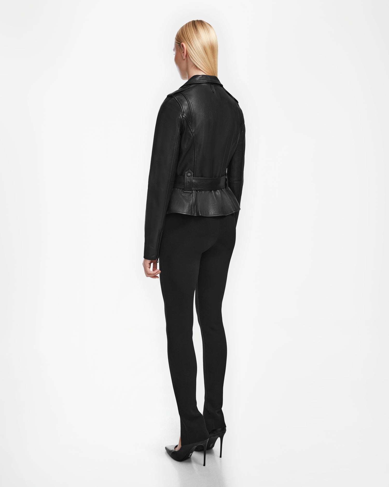 A woman with straight blonde hair faces away, wearing the Rudsak YEVA - 8124511 Black leather jacket, black fitted pants, and black high heels against a plain white background.