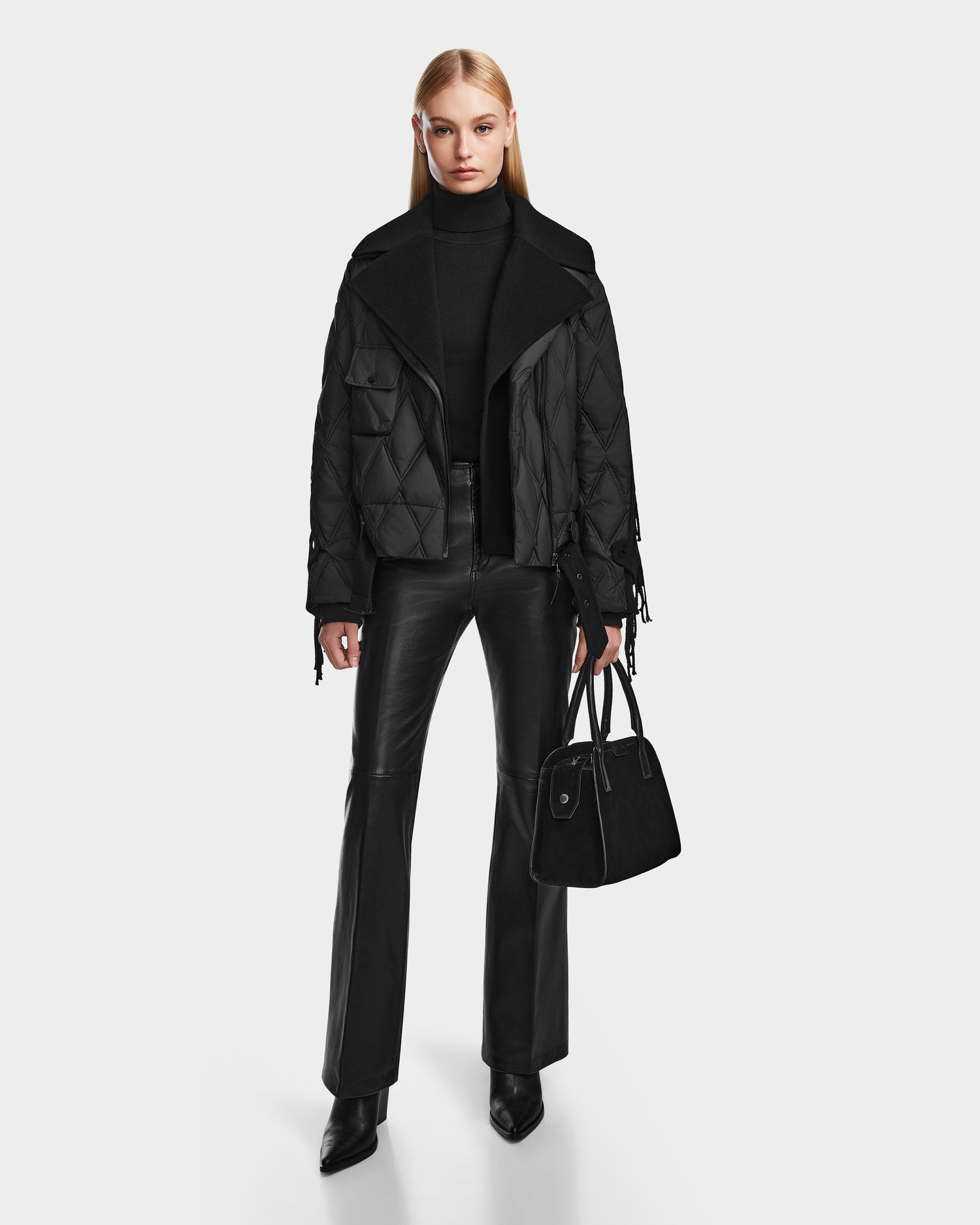 A woman with straight blonde hair faces forward wearing the Rudsak ZOLA - 8124712 Black quilted down jacket, black turtleneck, leather pants, heeled boots, and holding a black handbag against a plain white background.