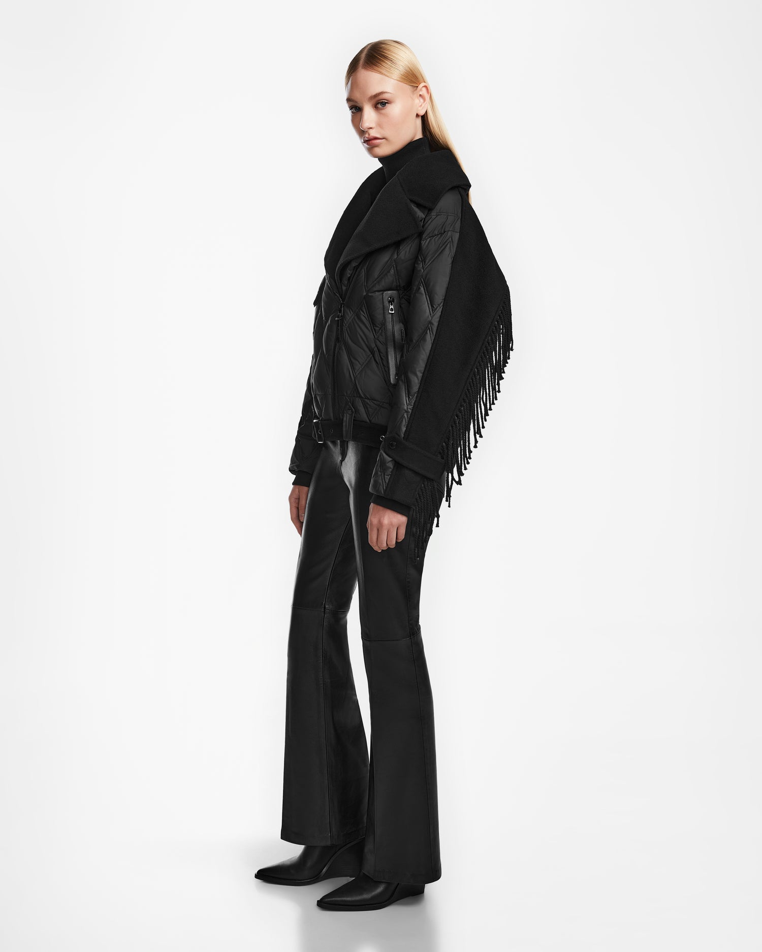 A woman with long blonde hair models the Rudsak ZOLA - 8124712 Black quilted down jacket with an oversized collar and fringed scarf, paired with black flared pants and boots, against a white background.