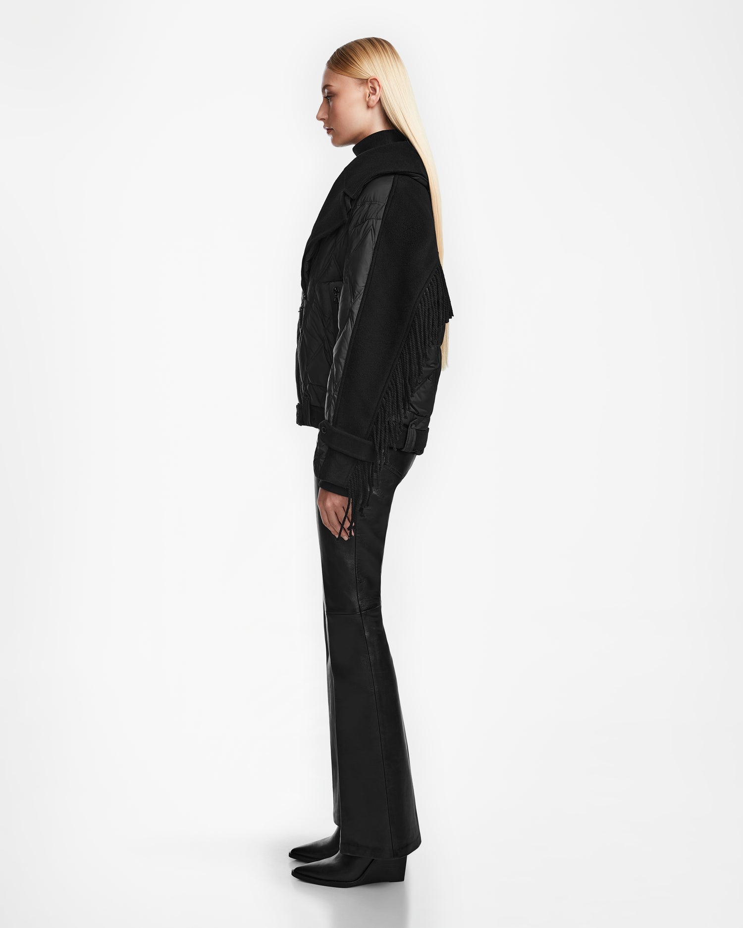 A woman with long blonde hair stands in profile against a white background, wearing the Rudsak ZOLA - 8124712 Black jacket, paired with black pants and black boots.