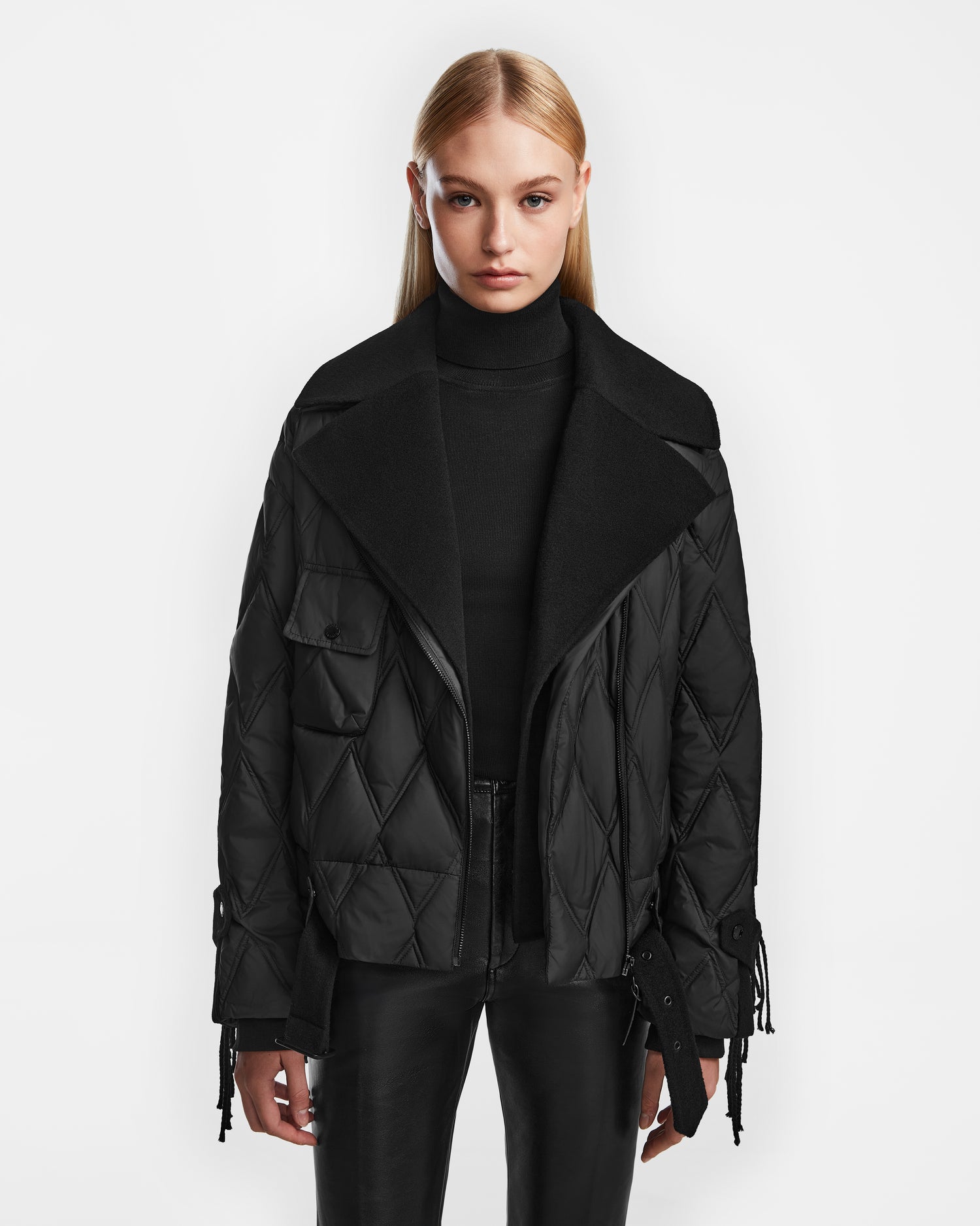 A woman with straight blonde hair wears the Rudsak ZOLA - 8124712 Black women's biker jacket over a black turtleneck and black leather pants, standing against a plain white background.