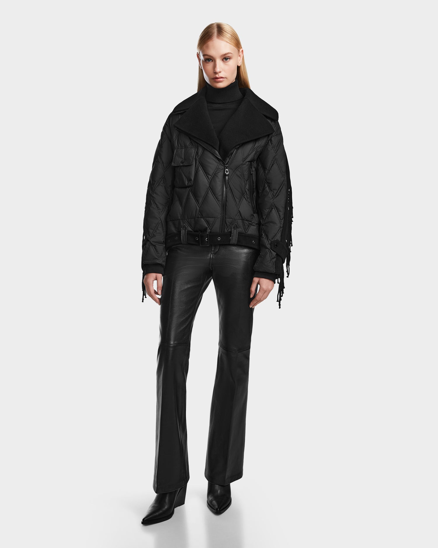 A person with long blonde hair wears the Rudsak ZOLA - 8124712 Black quilted down jacket, black turtleneck, leather pants, and boots, standing against a plain white background.