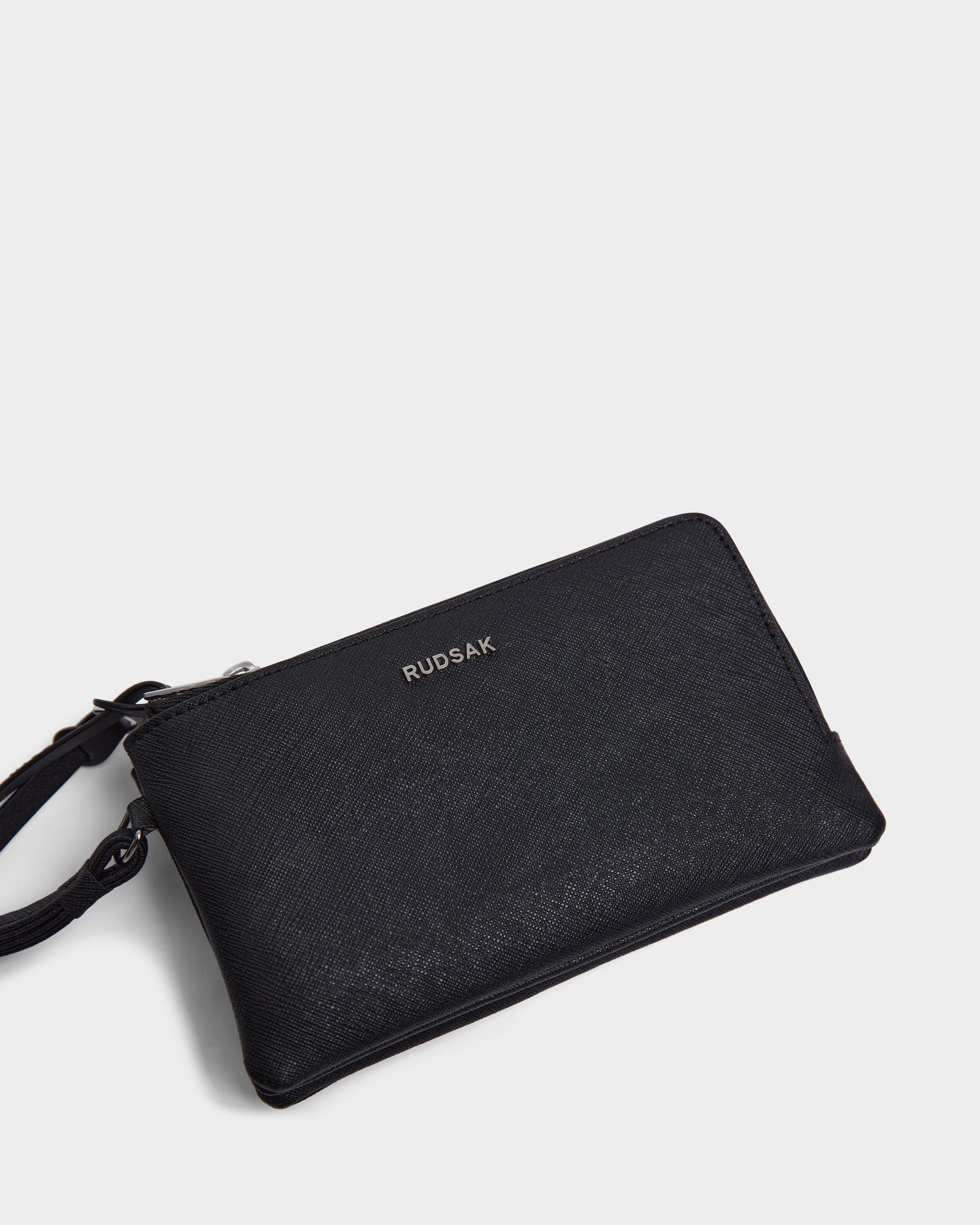The Rudsak ZURIE - L424501 Black is a womens textured leather wristlet wallet with a compact design, small strap, and silver RUDSAK logo on the front, shown against a plain white background.