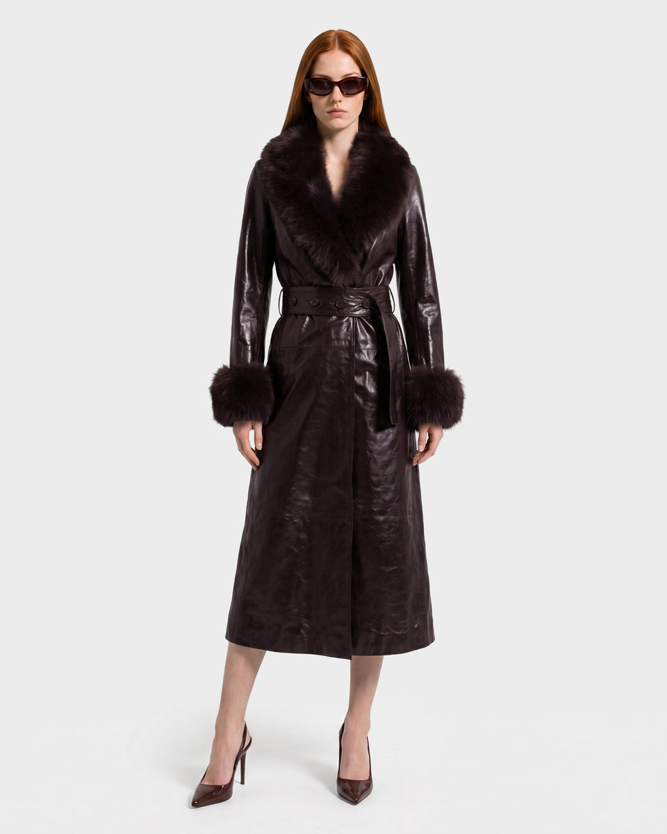 A woman with straight red hair wears dark sunglasses and the VIVIANNE - 8126933 Espresso, a midi-length dark brown leather trench from Rudsak featuring a faux fur collar and cuffs. She stands facing forward in pointed heels against a plain background.