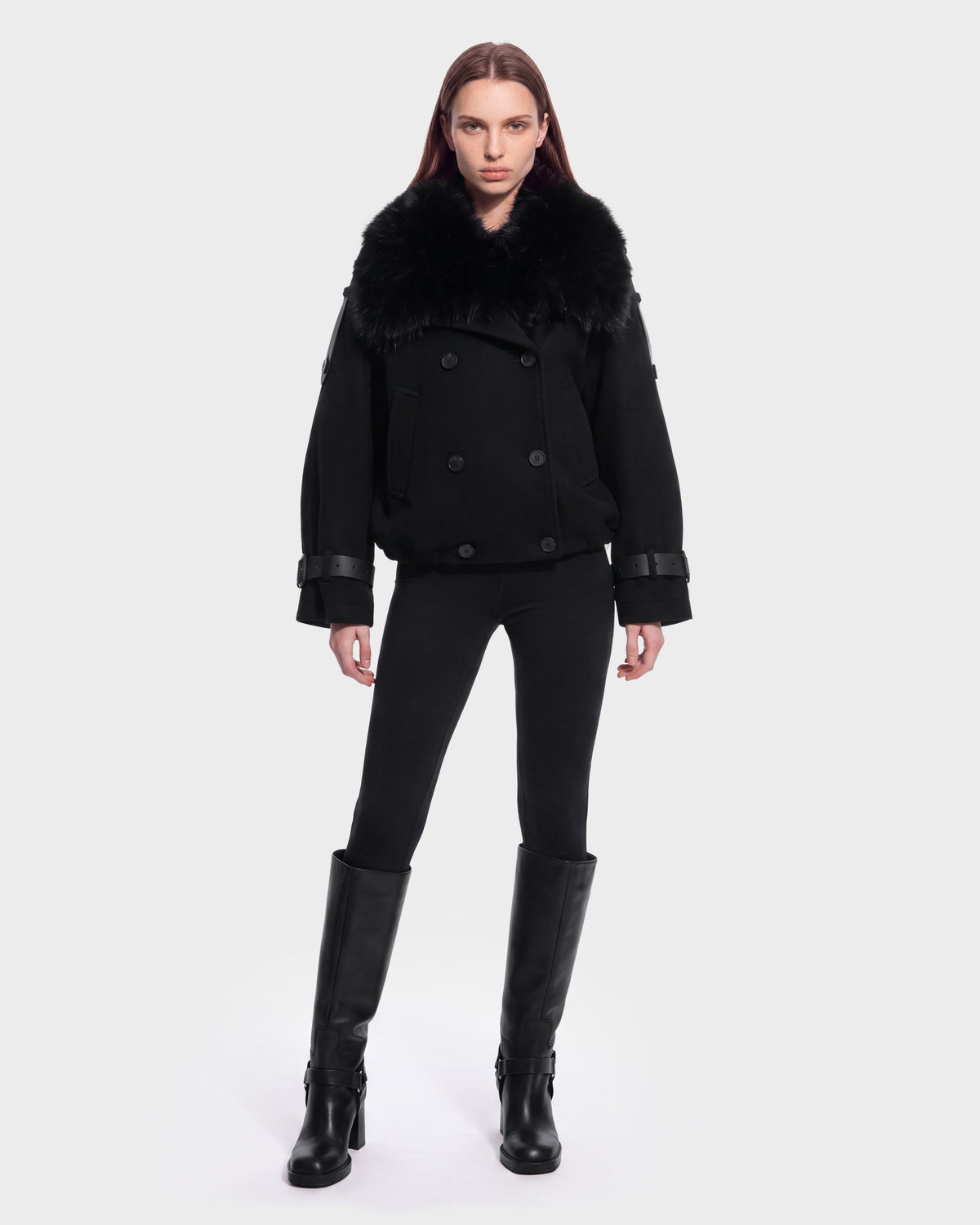 A person stands facing forward, wearing the Rudsak MAEMI - 8126985 Black luxury bomber jacket with a large removable faux fur collar, paired with black leggings and knee-high black boots against a plain white background.