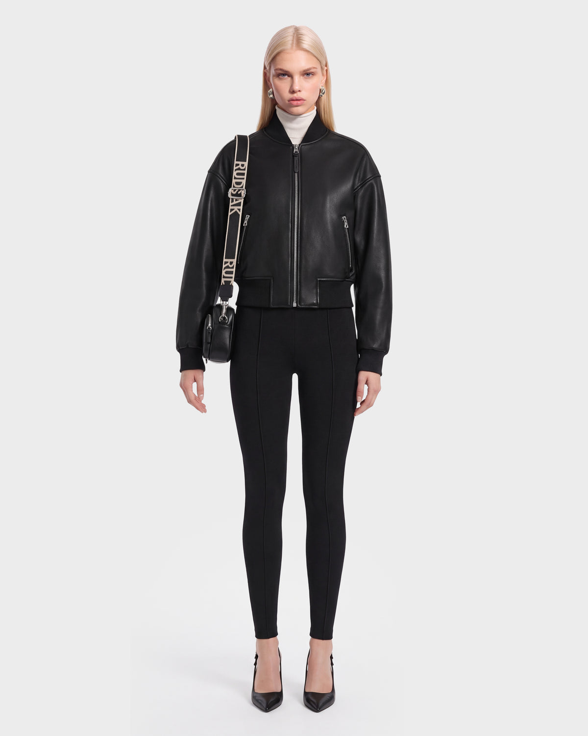 A woman stands facing forward against a plain light background, wearing the Rudsak EMERSON - 8125041 Black leather bomber jacket with a cropped silhouette, paired with black fitted pants, black heels, and a crossbody bag with a logo strap, showcasing contemporary outerwear style.