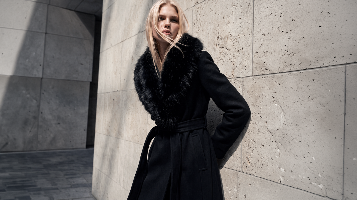 A blonde model wearing a black belted coat with a black fur collar, leaning against a textured concrete wall.