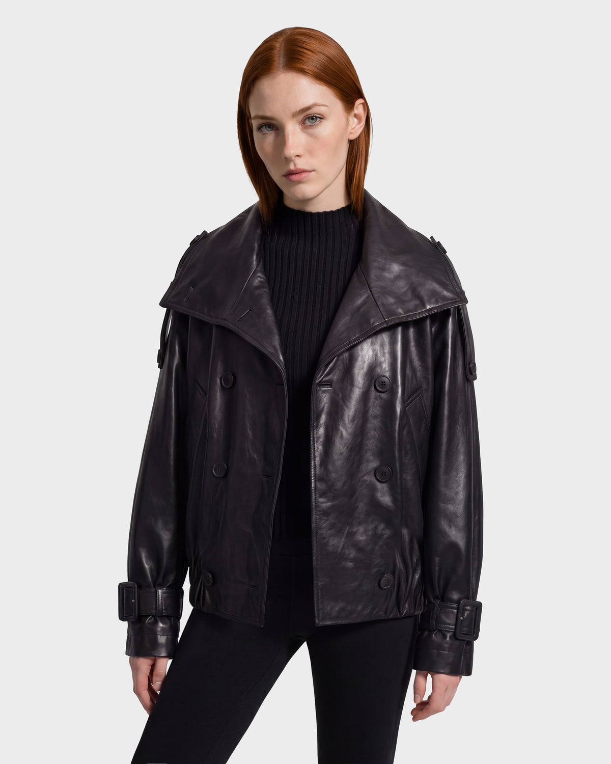 A woman wears a modern silhouette black JANELLE - 8126936 Black leather double-breasted jacket with a wide collar and buckled cuffs.