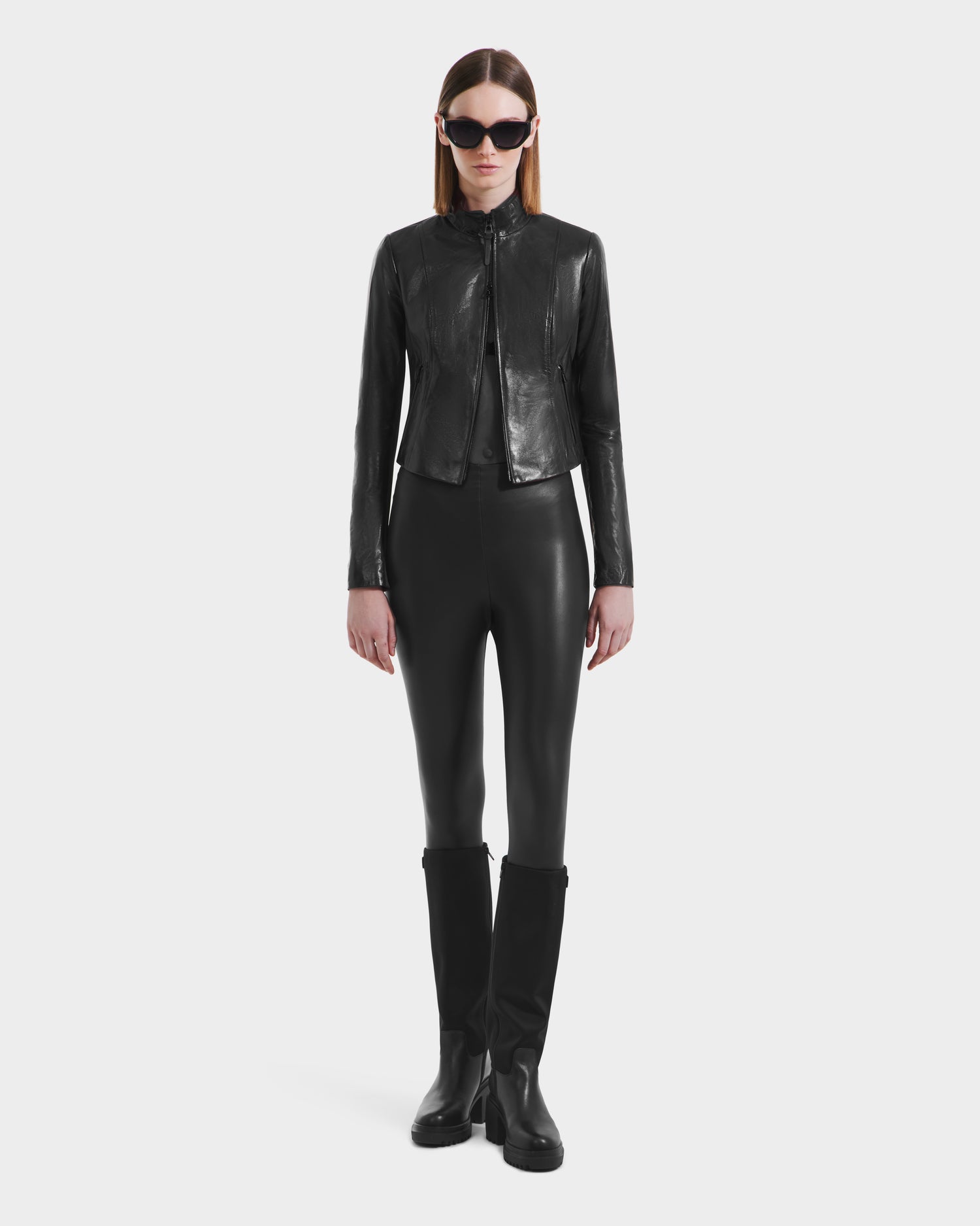 A person in black sunglasses, Rudsak's ABIGAIL N - 8125970 Black women's leather racer jacket, black leggings, and knee-high boots stands facing forward against a plain white background.