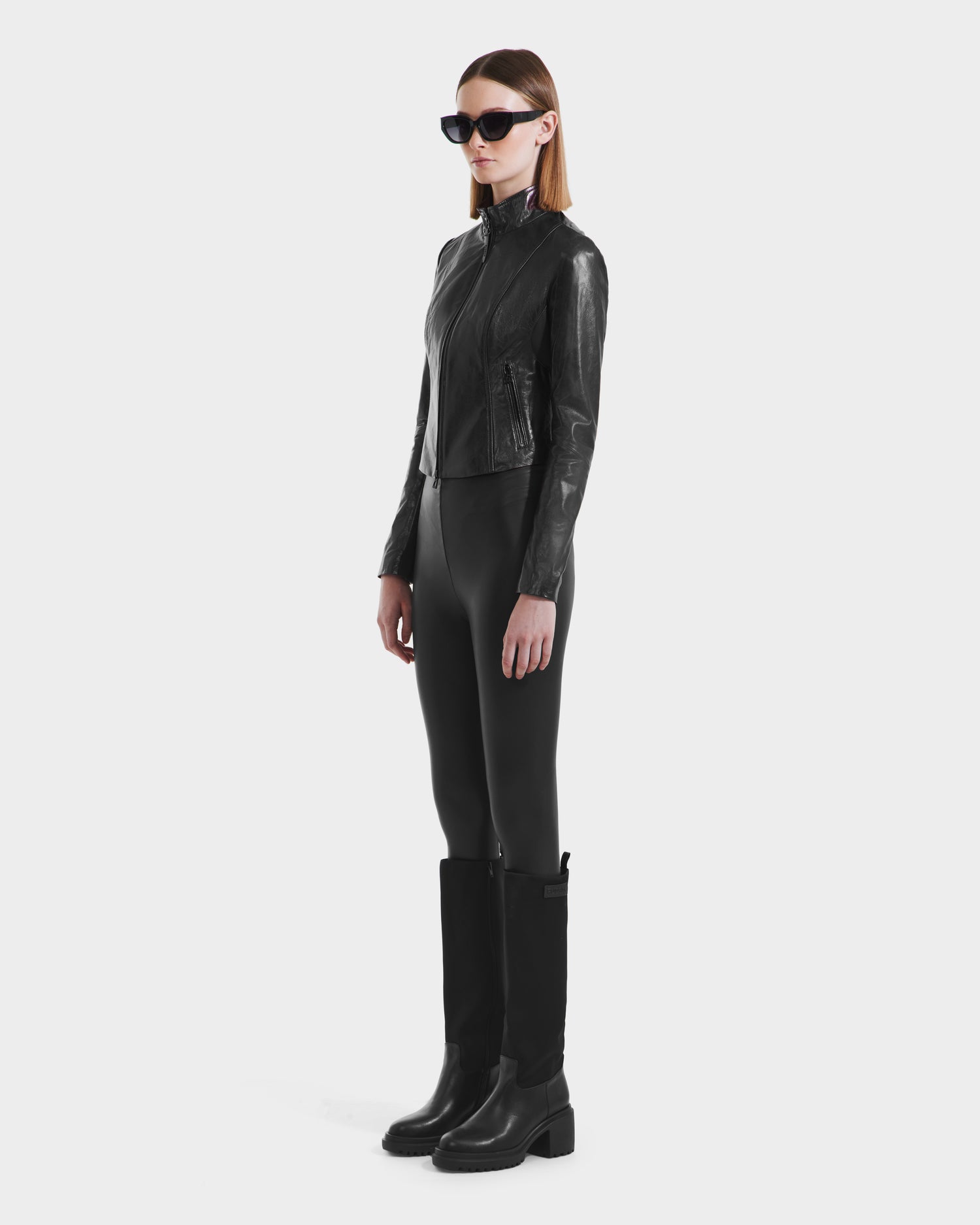 A woman stands against a plain white background wearing black sunglasses, the Rudsak ABIGAIL N - 8125970 Black fitted leather jacket, black fitted pants, and knee-high black boots.