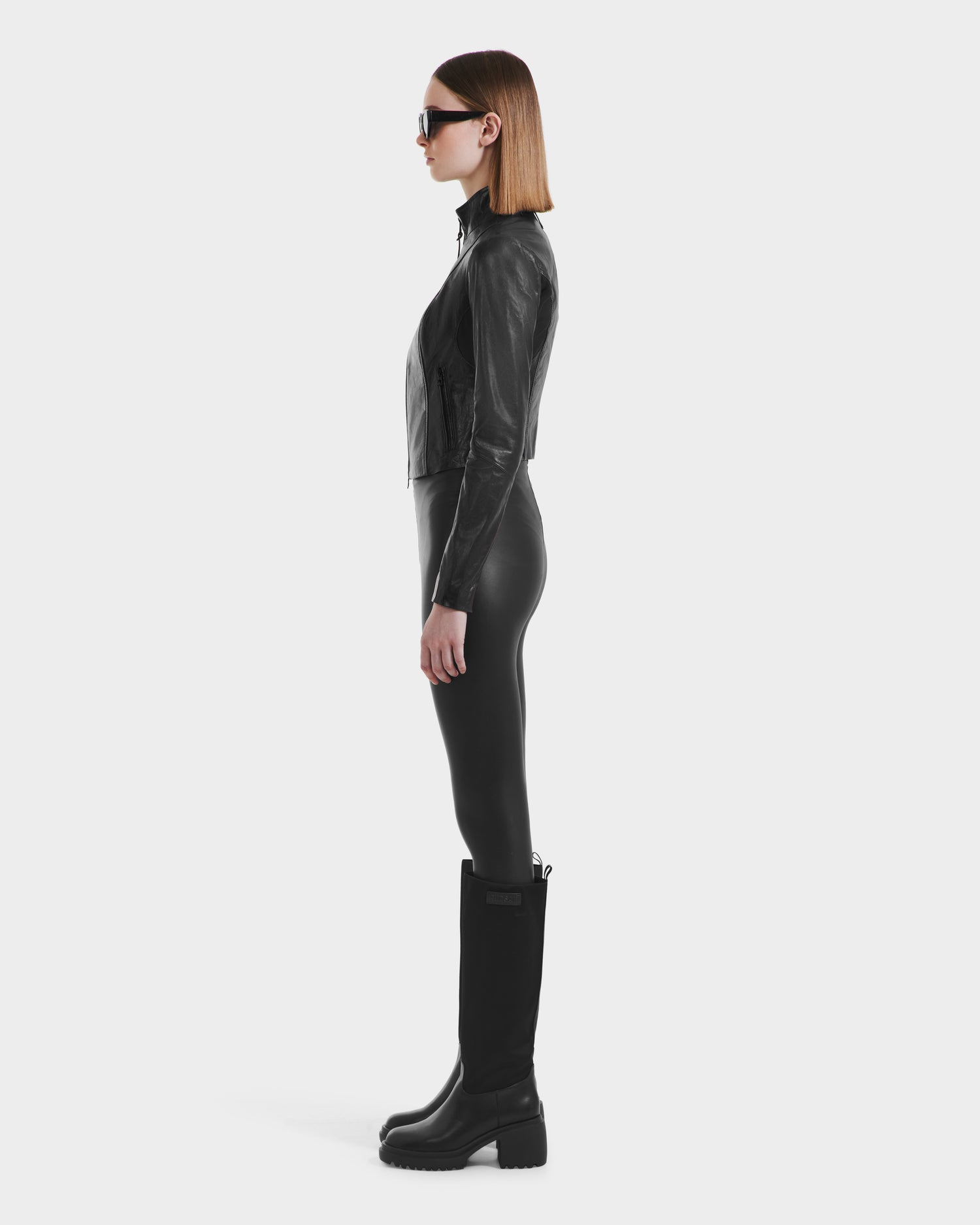 A person with straight, shoulder-length hair stands in profile wearing black sunglasses and the Rudsak ABIGAIL N - 8125970 Black women's leather racer jacket, styled with fitted pants and tall boots against a plain white background.