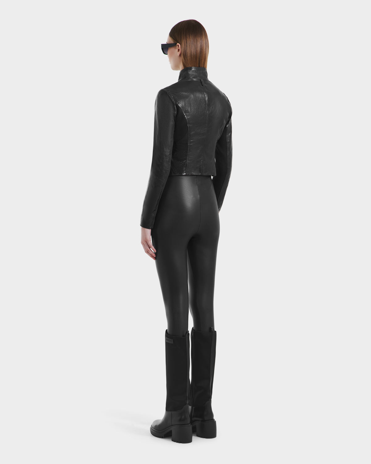 A person wearing black sunglasses, black pants, boots, and a Rudsak ABIGAIL N - 8125970 Black fitted leather jacket stands facing away from the camera against a plain white background.
