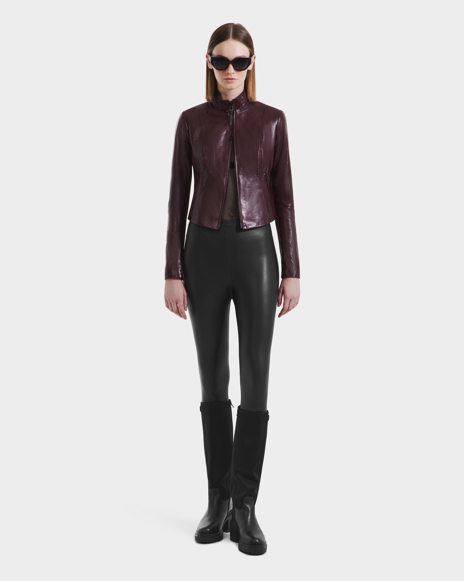 A woman faces forward wearing the Rudsak ABIGAIL - 8124560 Bordeaux leather jacket, paired with black leather pants, black knee-high boots, and black sunglasses against a plain white background.
