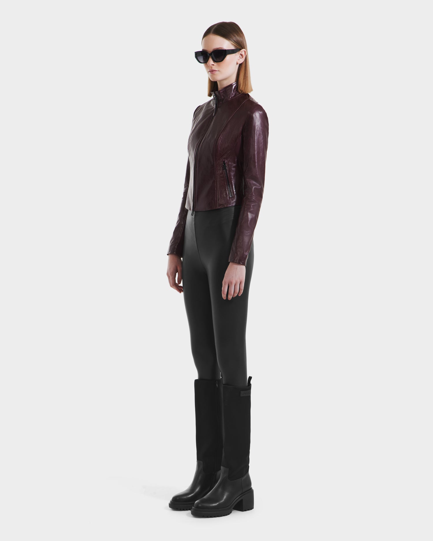 A woman in a RUDSAK ABIGAIL - 8124560 Bordeaux leather racer jacket, black leggings, tall black boots, and black sunglasses stands against a plain white background.