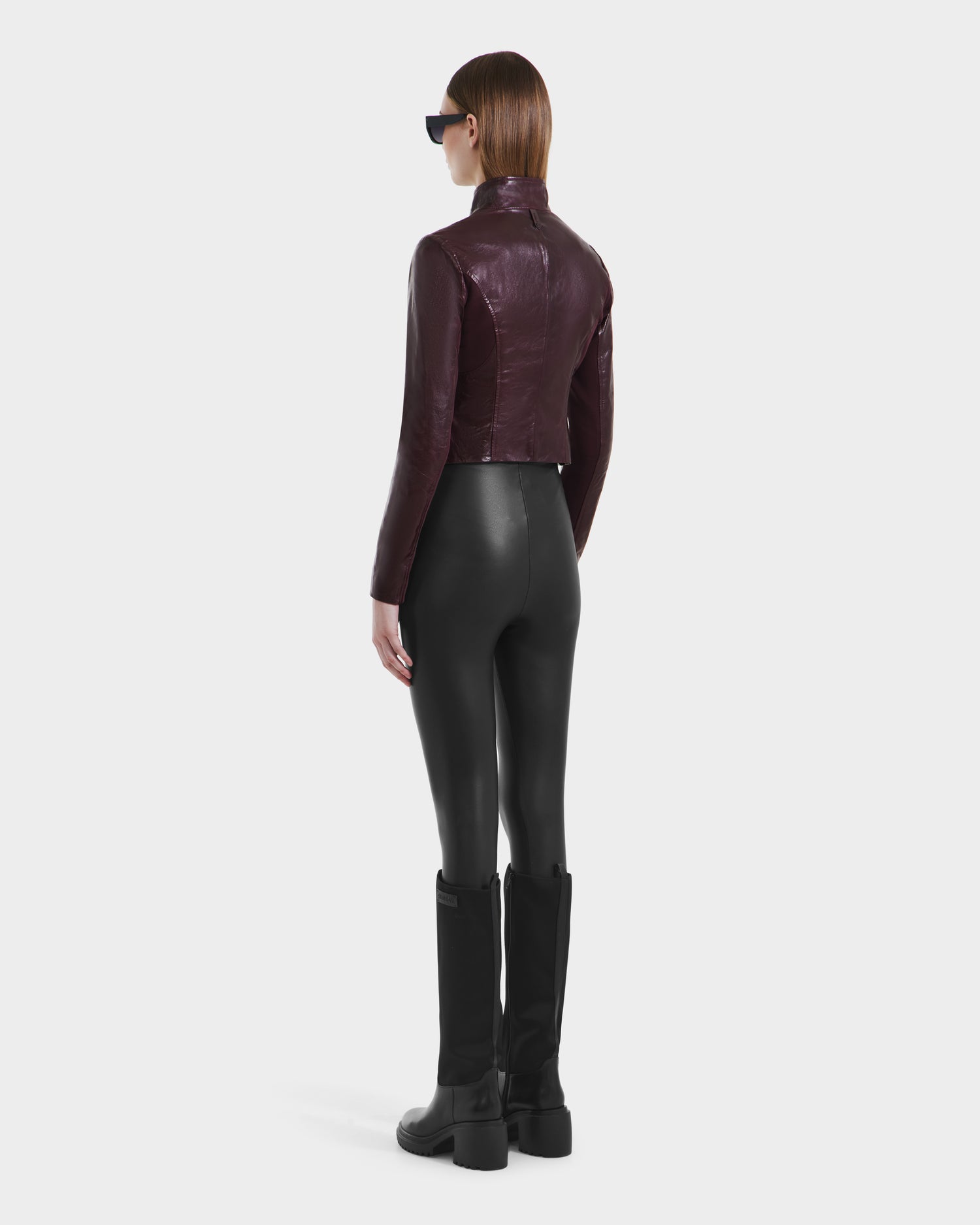 A person with straight brown hair, wearing sunglasses and a fitted Rudsak ABIGAIL - 8124560 Bordeaux leather jacket, black leggings, and black knee-high boots stands facing away against a plain white background.