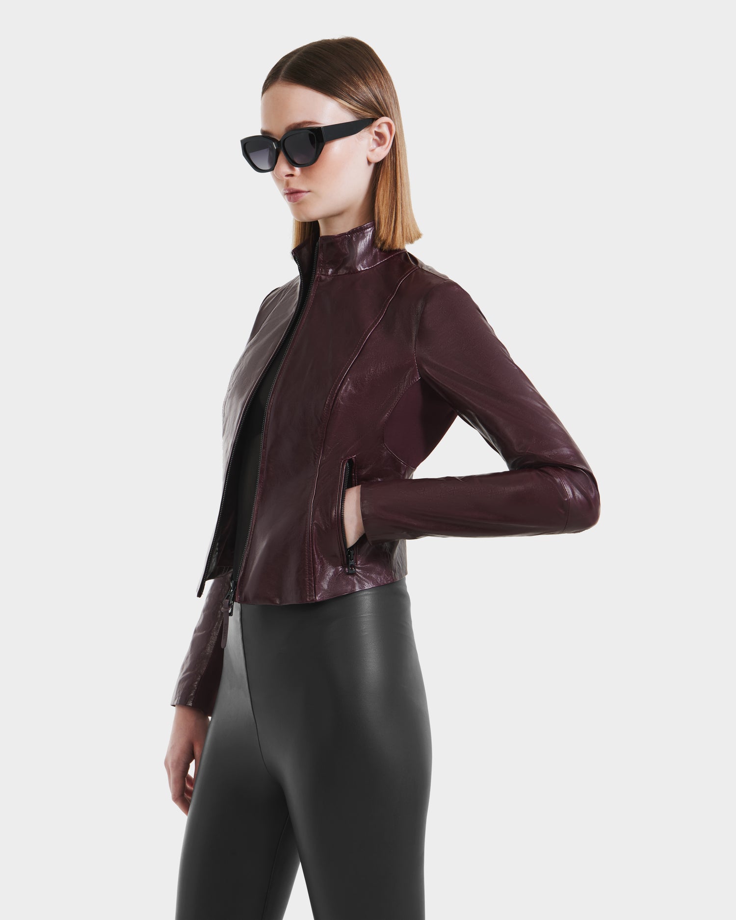 A woman in dark sunglasses, the ABIGAIL - 8124560 Bordeaux leather racer jacket by Rudsak, and black high-waisted pants stands against a plain white background with one hand in her jacket pocket.