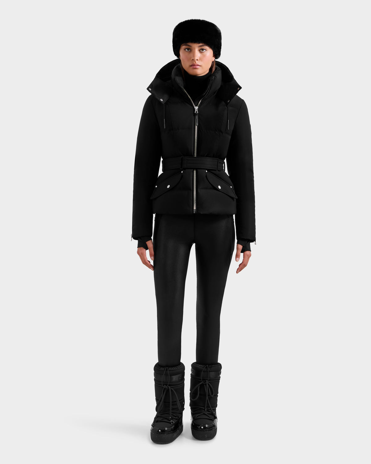 A woman wearing the Rudsak ADELINE H - 8125587 Black belted down puffer jacket, with a black fur hat, turtleneck, fitted pants, and winter boots stands facing forward against a plain white background.