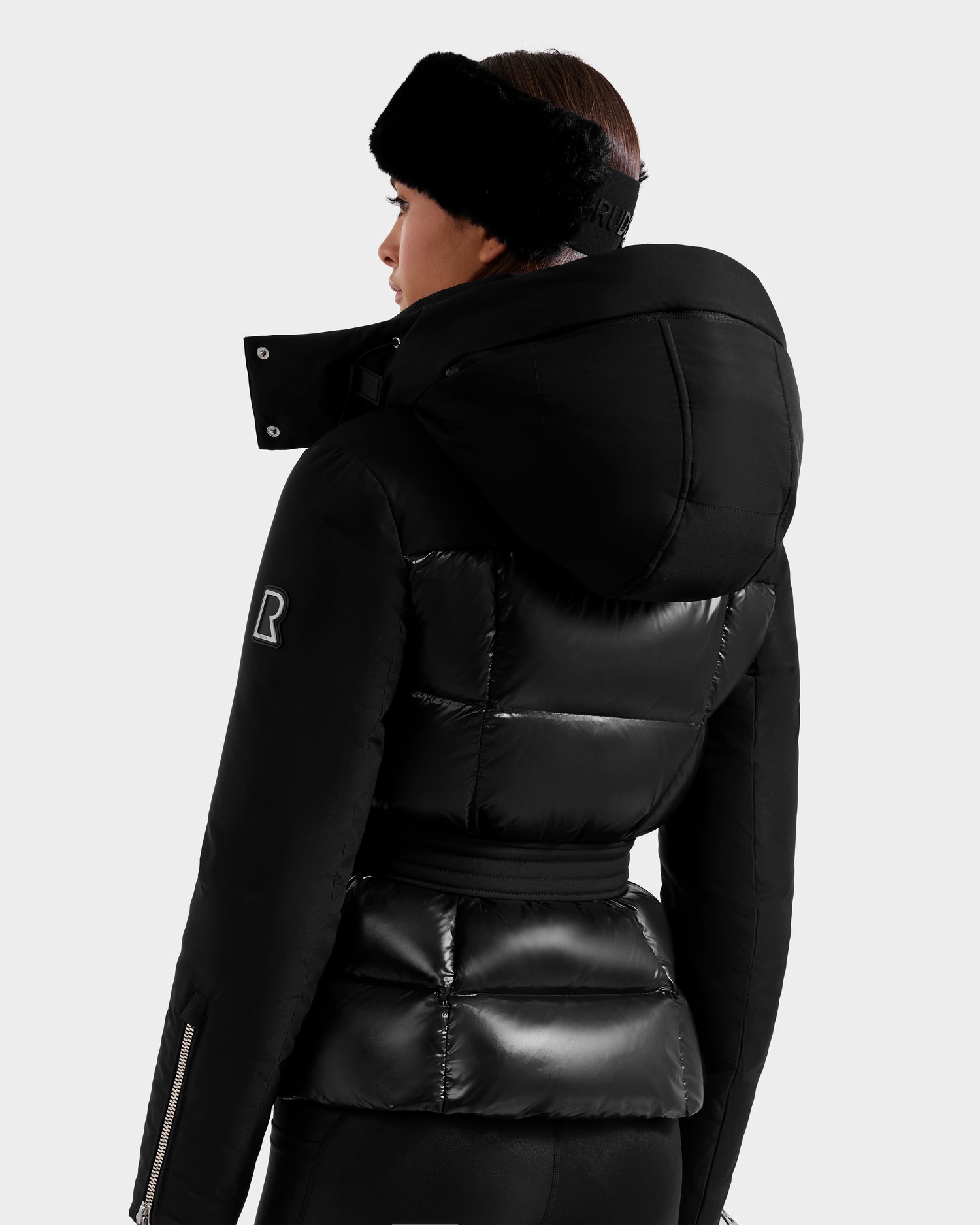A woman is seen from behind wearing the Rudsak ADELINE H - 8125587 black down puffer, featuring a shiny finish, R logo on the sleeve, hood up, and paired with black pants against a plain light background.