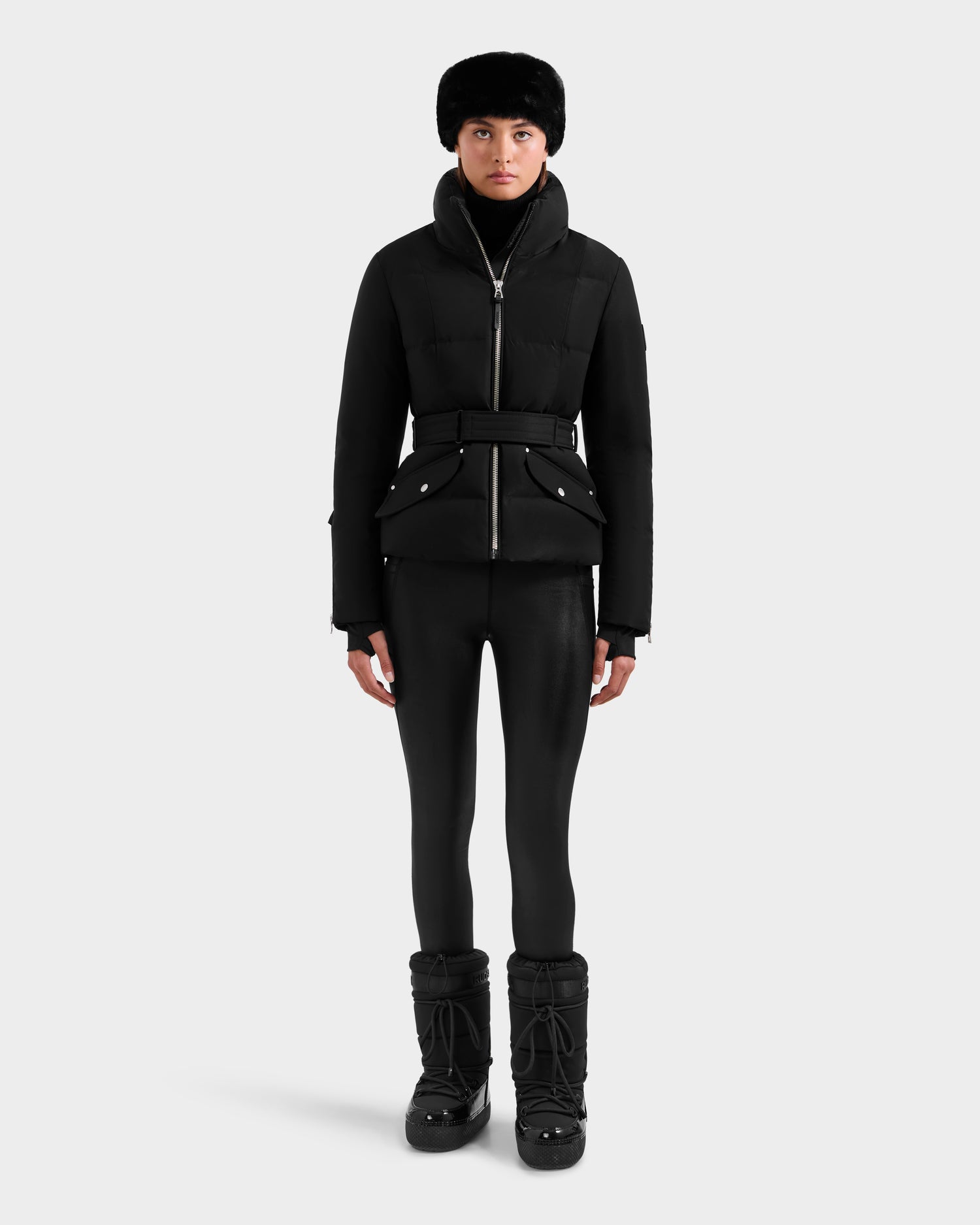 A person stands facing forward, wearing a black Rudsak ADELINE H - 8125587 jacket belted at the waist, black winter hat, leggings, and boots against a plain light gray background.
