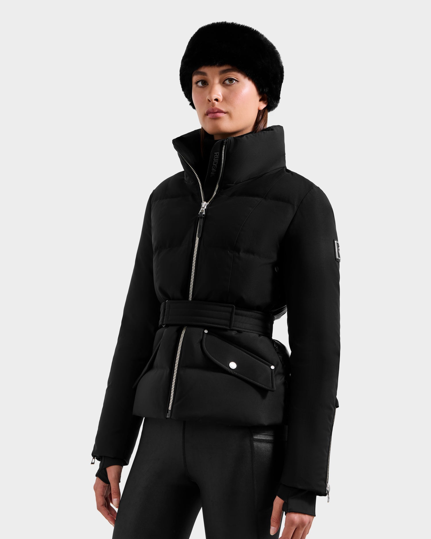 A woman stands against a plain light gray background, wearing the Rudsak ADELINE H - 8125587 Black women’s down puffer jacket with a belt, black leather pants, and a black fur hat. She has a neutral expression and her hands by her sides.