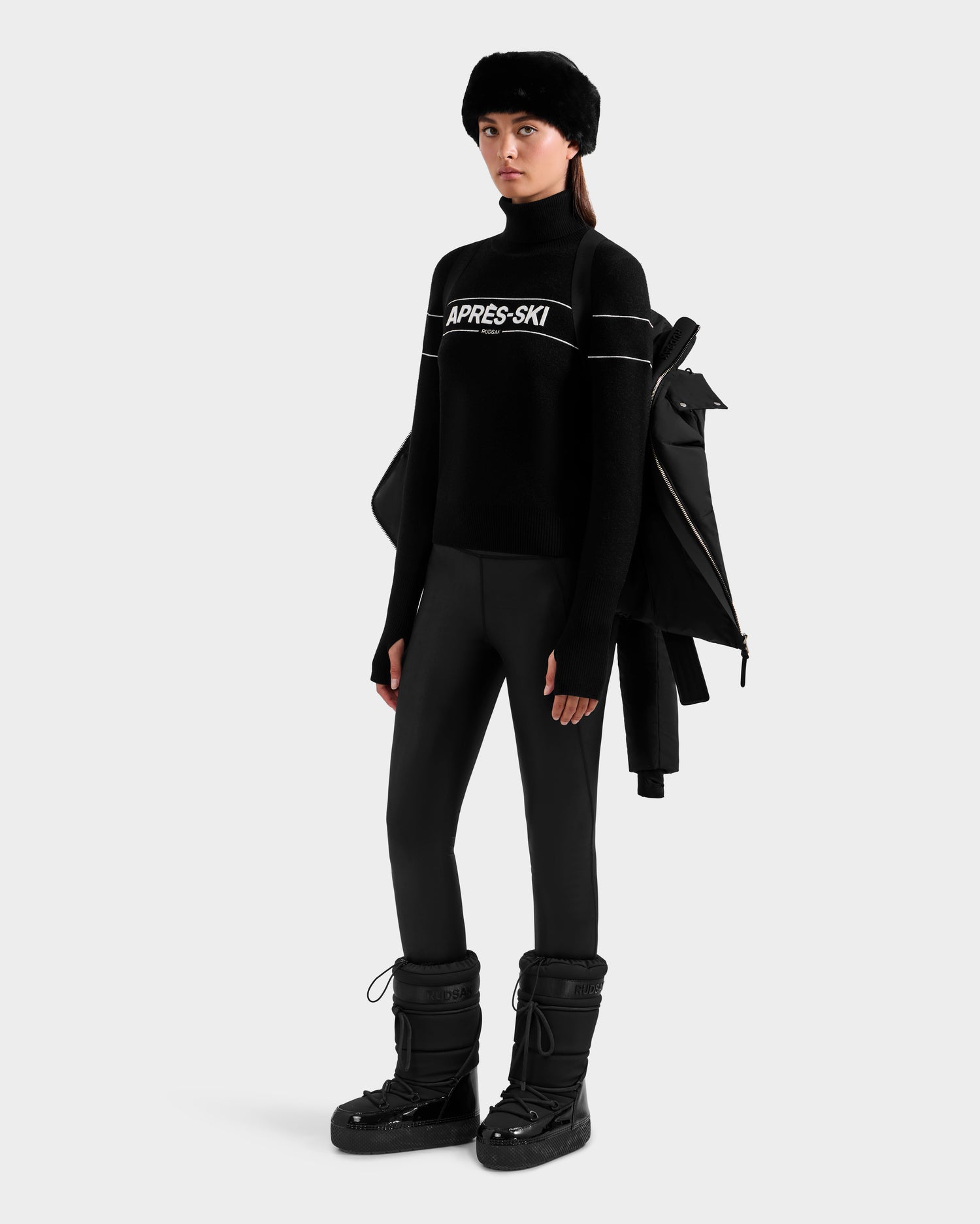 A person wearing a black fur hat, black APRÈS-SKI turtleneck sweater, black leggings, black snow boots, and carrying a Rudsak ADELINE H - 8125587 Black jacket over their shoulder stands against a plain background.