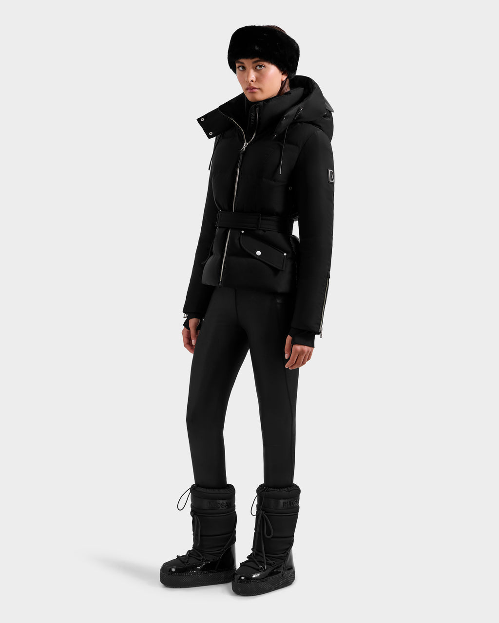 A person models the Rudsak ADELINE H - 8125587 Black, a stylish women’s down puffer winter coat, paired with fitted pants, snow boots, and a fur hat against a plain white background.
