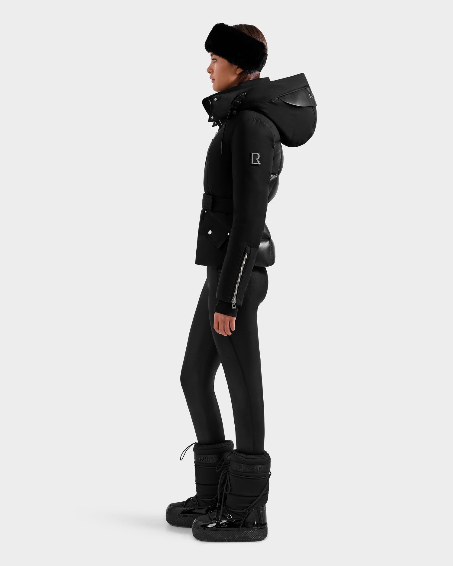 A woman in the Rudsak ADELINE H - 8125587 Black down puffer with a large hood, black leggings, winter boots, and a black headband stands in profile against a plain light background.