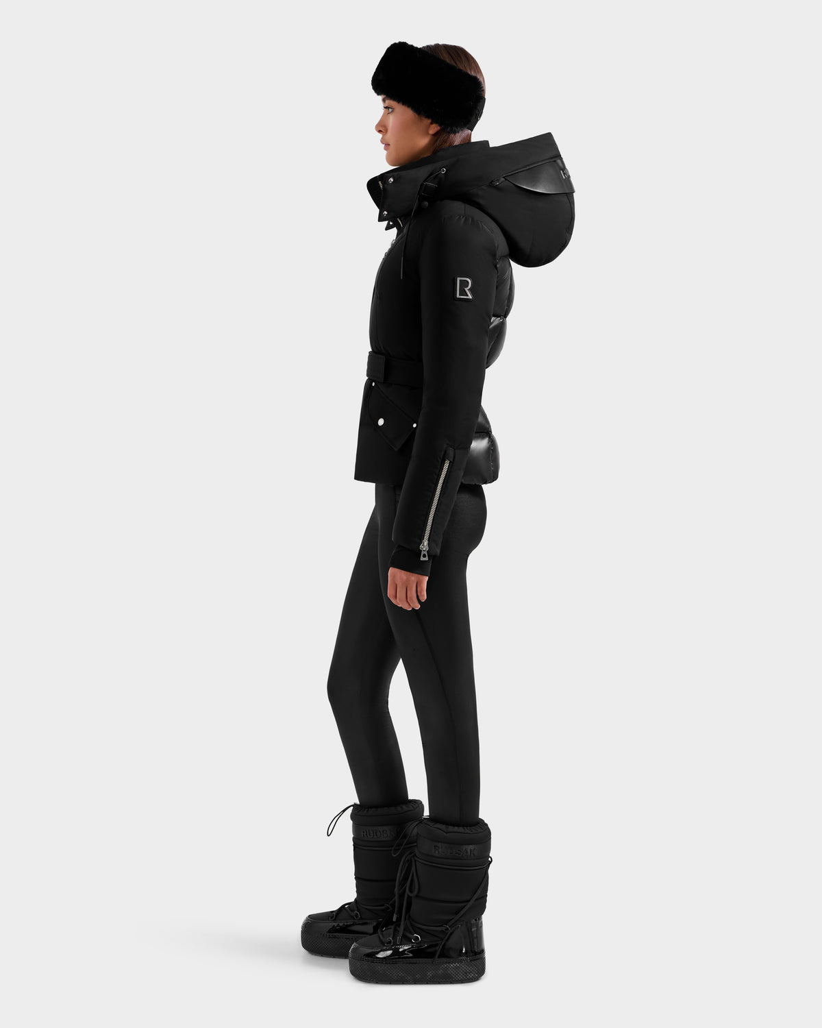 A woman in the Rudsak ADELINE H - 8125587 Black down puffer with a large hood, black leggings, winter boots, and a black headband stands in profile against a plain light background.