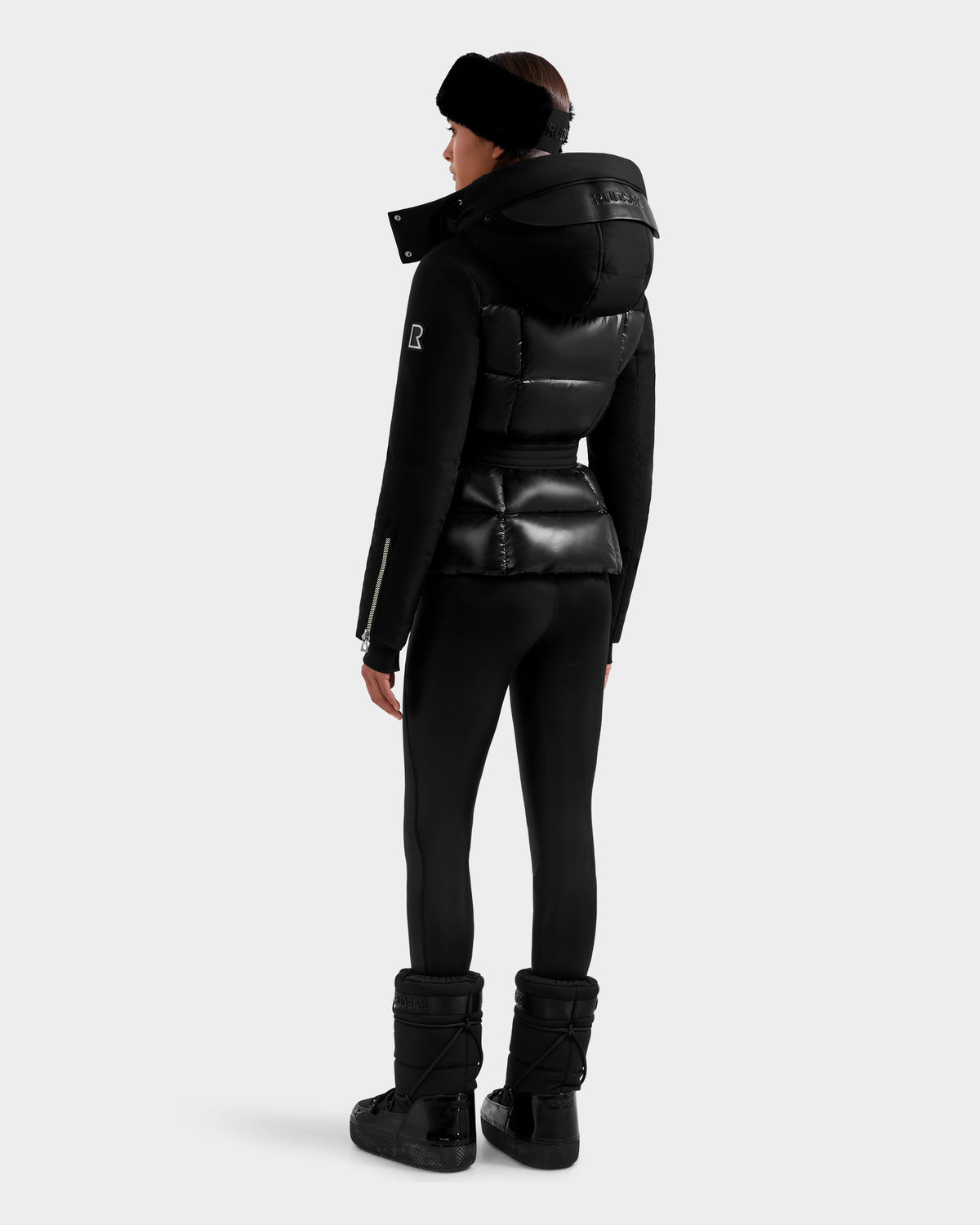 A person stands facing away, wearing the sleek Rudsak ADELINE H - 8125587 Black women’s down puffer with a hood, black leggings, boots, and a headband. The modern winter-ready look stands out against a plain light background.