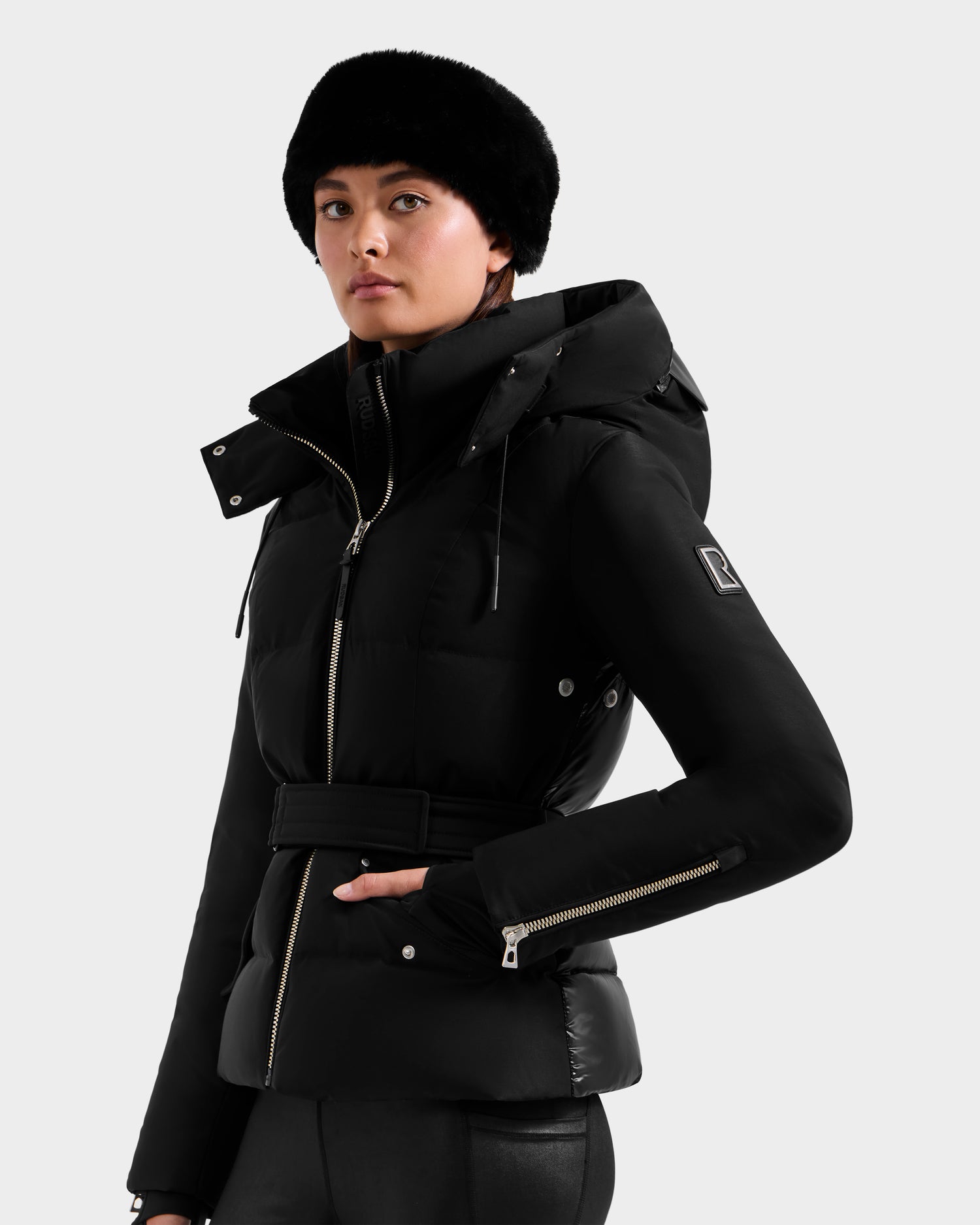 A woman wears the Rudsak ADELINE H - 8125587 Black belted women’s down puffer with a fur hat, zippered sleeves, and side pockets, standing against a plain light background.