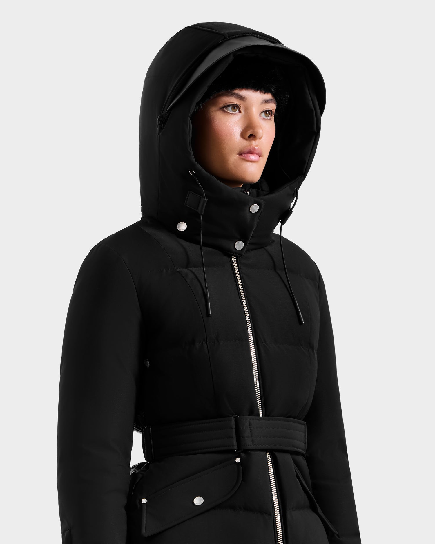 A person wearing the Rudsak ADELINE H - 8125587 black winter coat with a belted waist, silver zipper, and button details stands against a plain light background.
