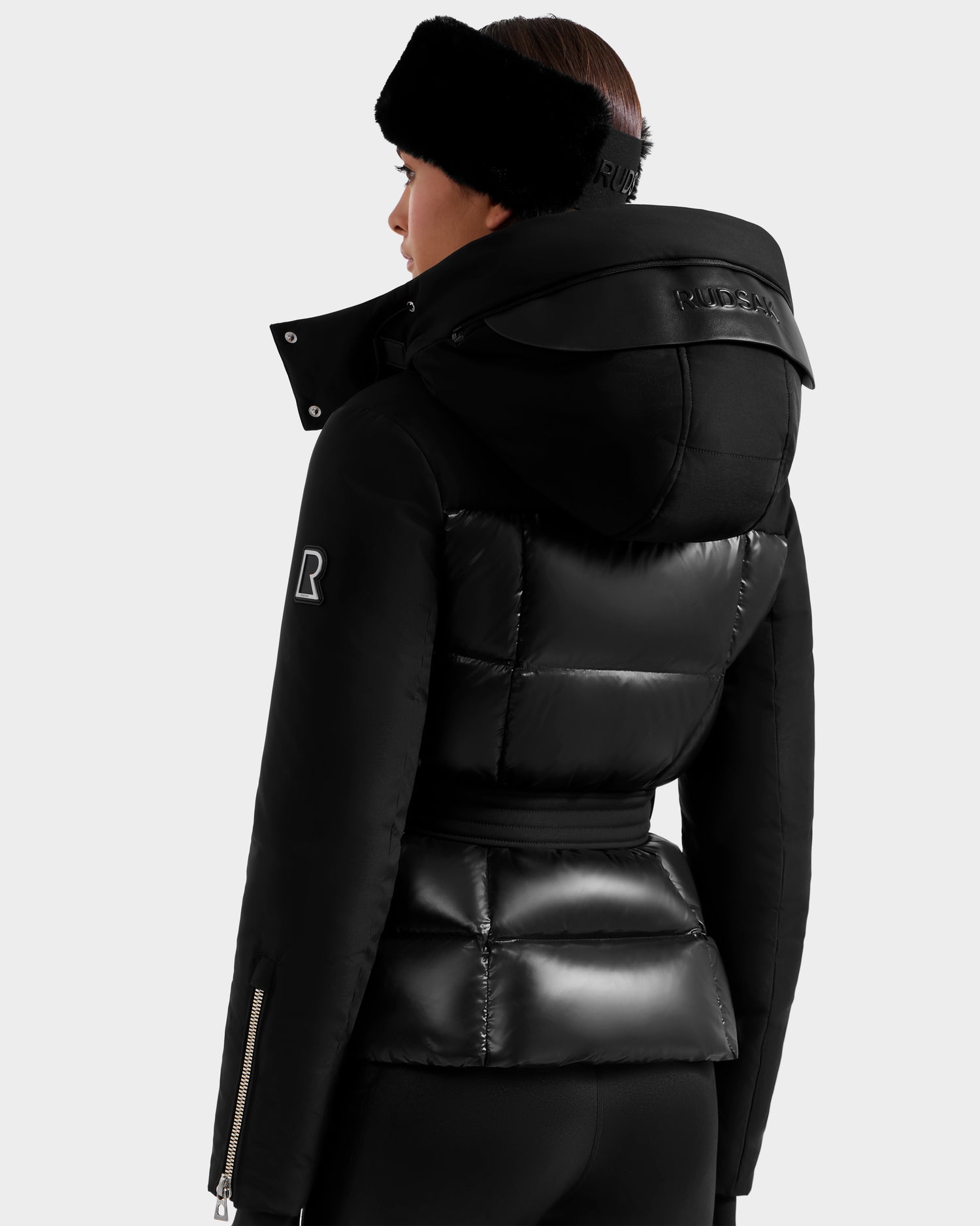 A woman facing away wears the Rudsak ADELINE H - 8125587 Black, a women's down puffer with glossy and matte panels, high collar, hood, and R logo on the sleeve, paired with a black headband and pants.