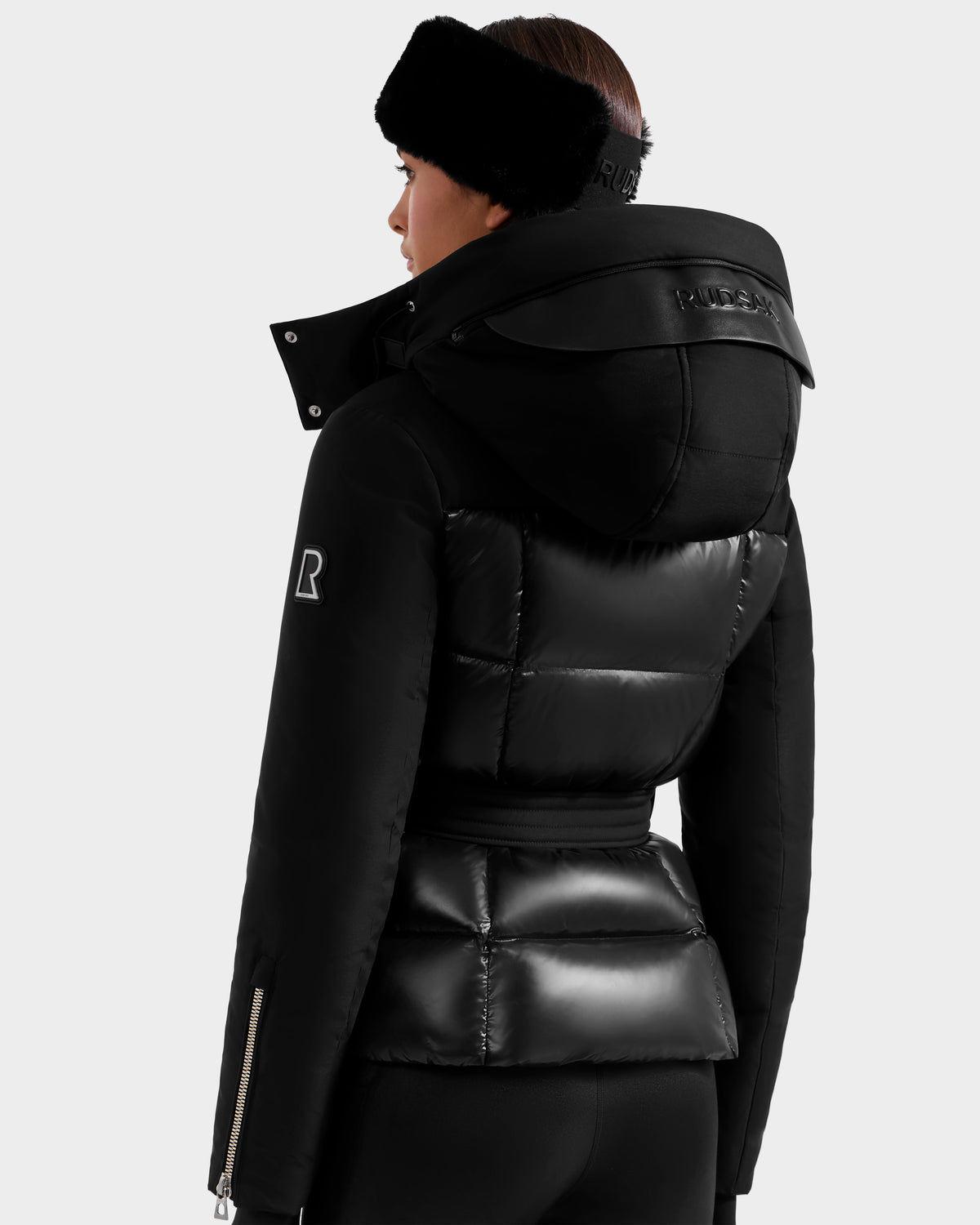 A woman facing away wears the Rudsak ADELINE H - 8125587 Black, a women's down puffer with glossy and matte panels, high collar, hood, and R logo on the sleeve, paired with a black headband and pants.