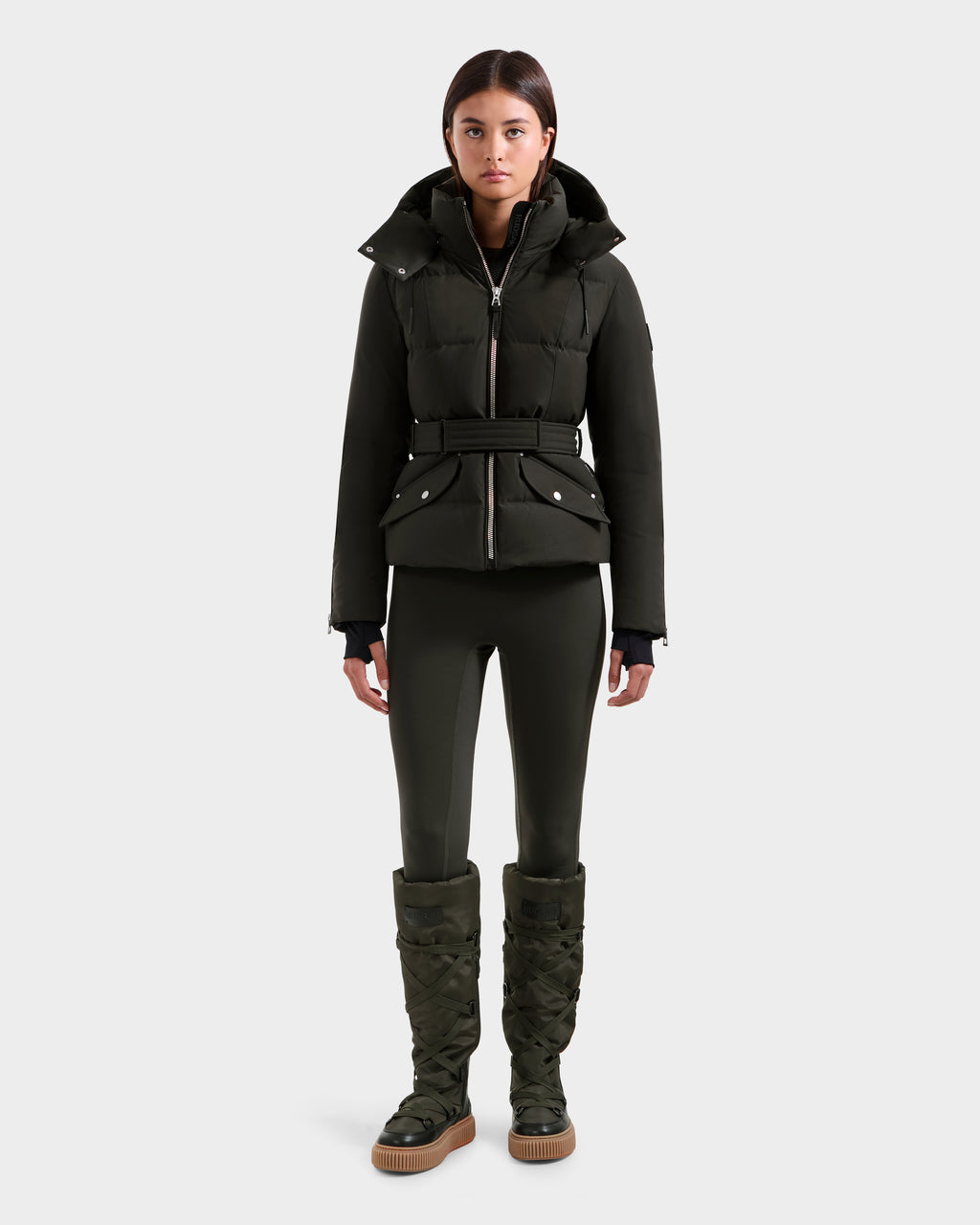 A woman faces forward in a Rudsak ADELINE H - 8125587 dark olive women's down puffer jacket with a belted waist, paired with matching fitted leggings and knee-high quilted boots against a plain light gray background.